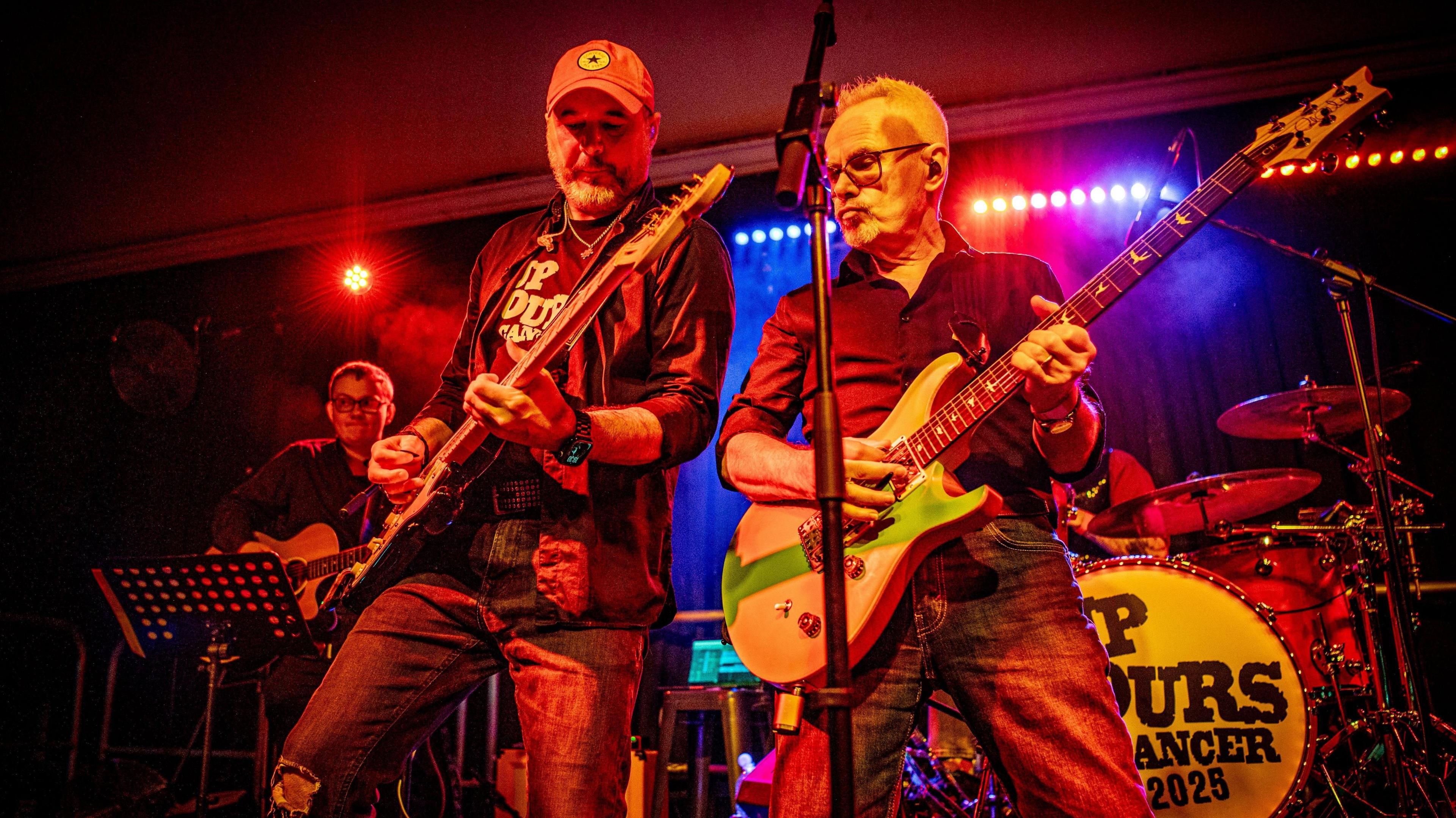 Two men on stage, Steve Young and Nik Kershaw. They are both holding guitars. In the background is another guitarist and a set of drums.