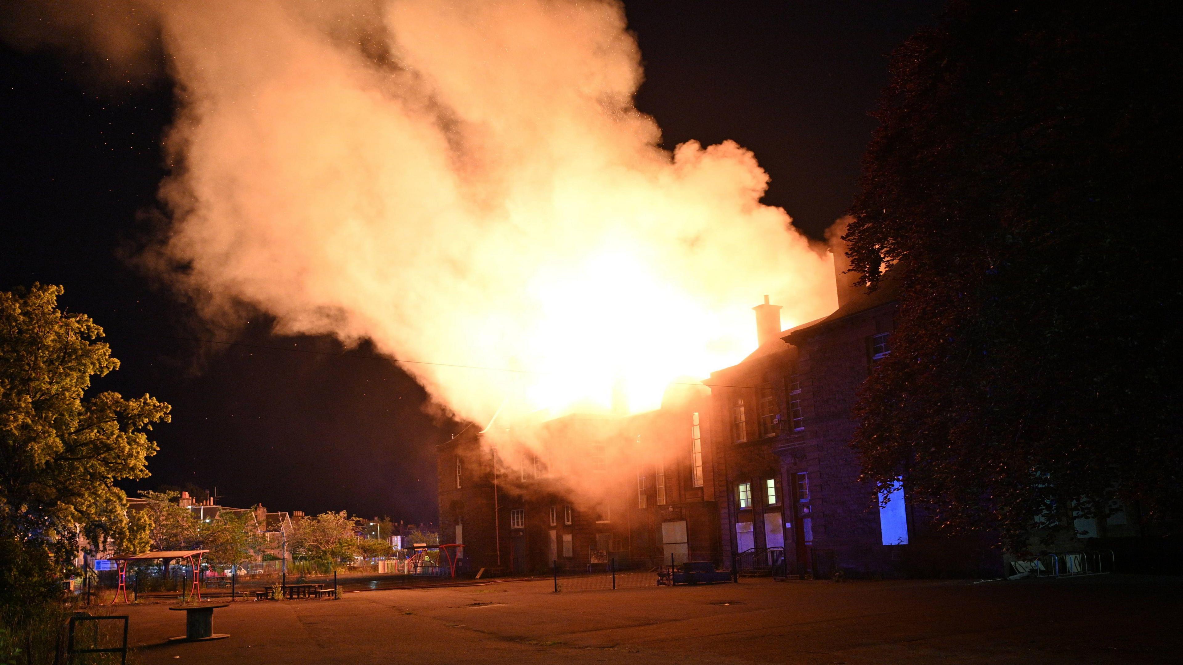 A large fire at a former primary school in Perth. The fire is bright orange and can be seen coming out of the top of the building, which is dark. The sky is also dark.
