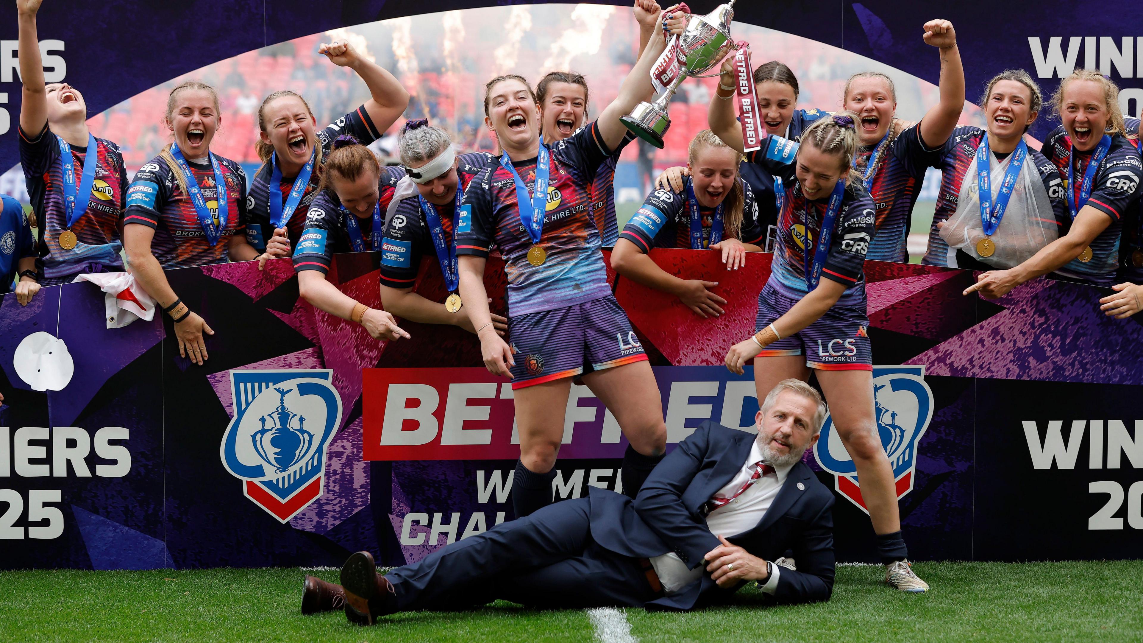 Wigan co-captains Vicky Molyneux and Georgia Wilson lift the trophy as head coach Denis Betts lies on the ground after the Rugby League Women's Challenge Cup Final between St Helens and Wigan Warriors at Wembley Stadium on June 7th 2025