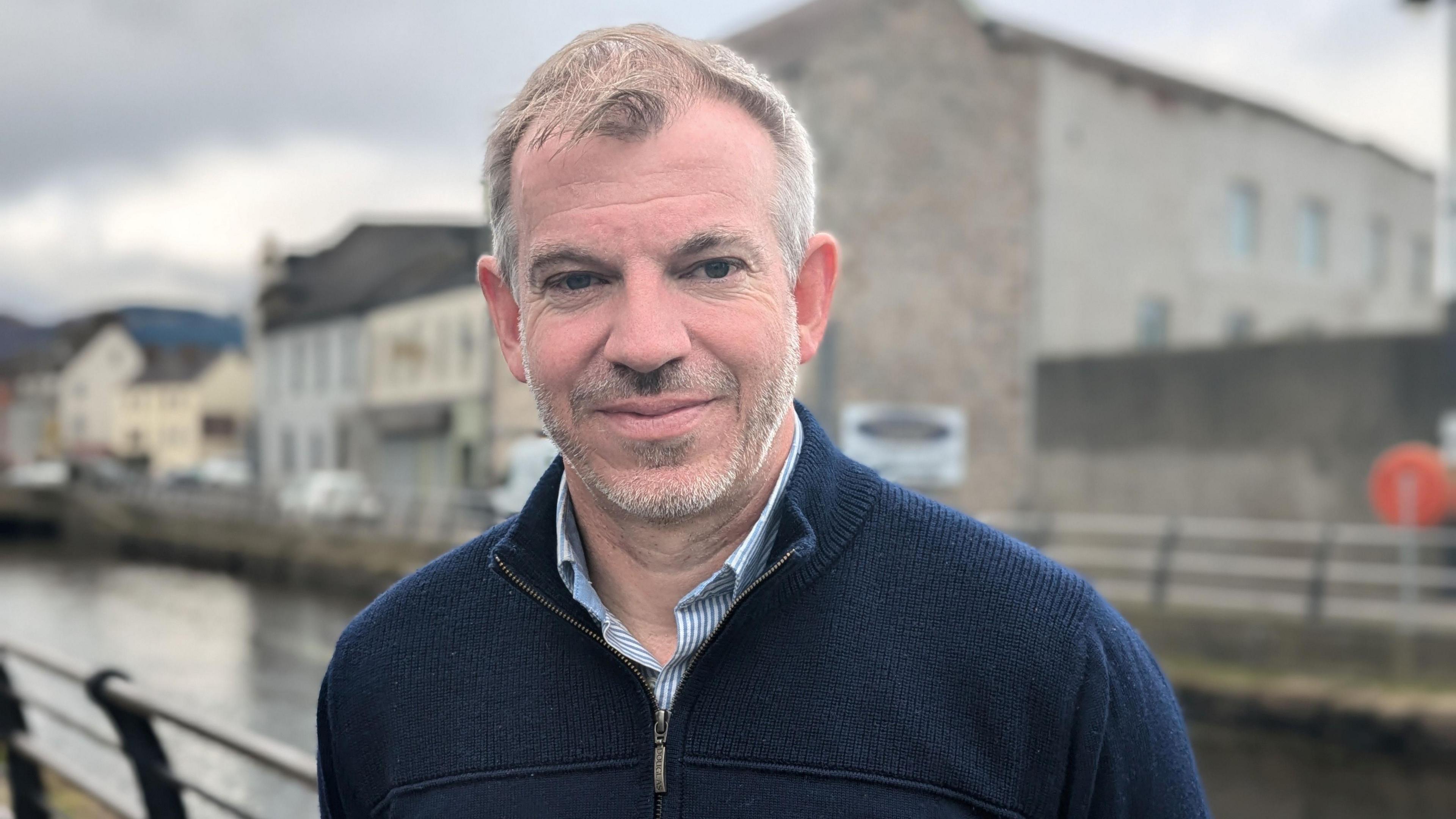 A man standing looking at the camera with a slight smile. Behind him there is a canal and some buildings.