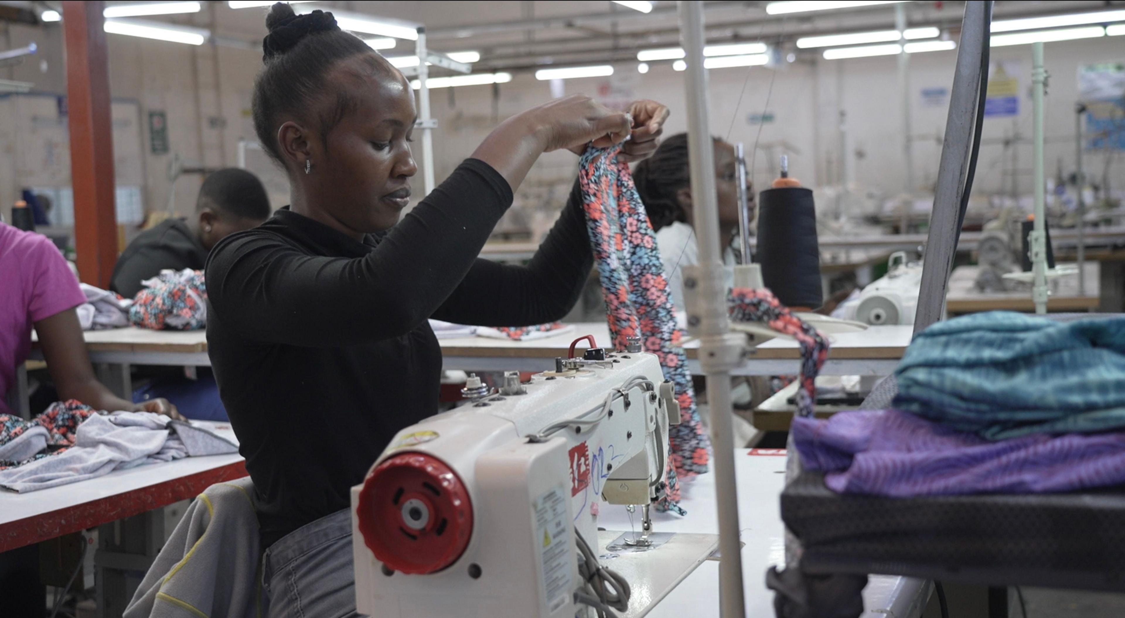 A worker sitting at a sewing machine inspecting a garment that she has just sewn. There are other sewers seated at machines around her on the factory floor. 