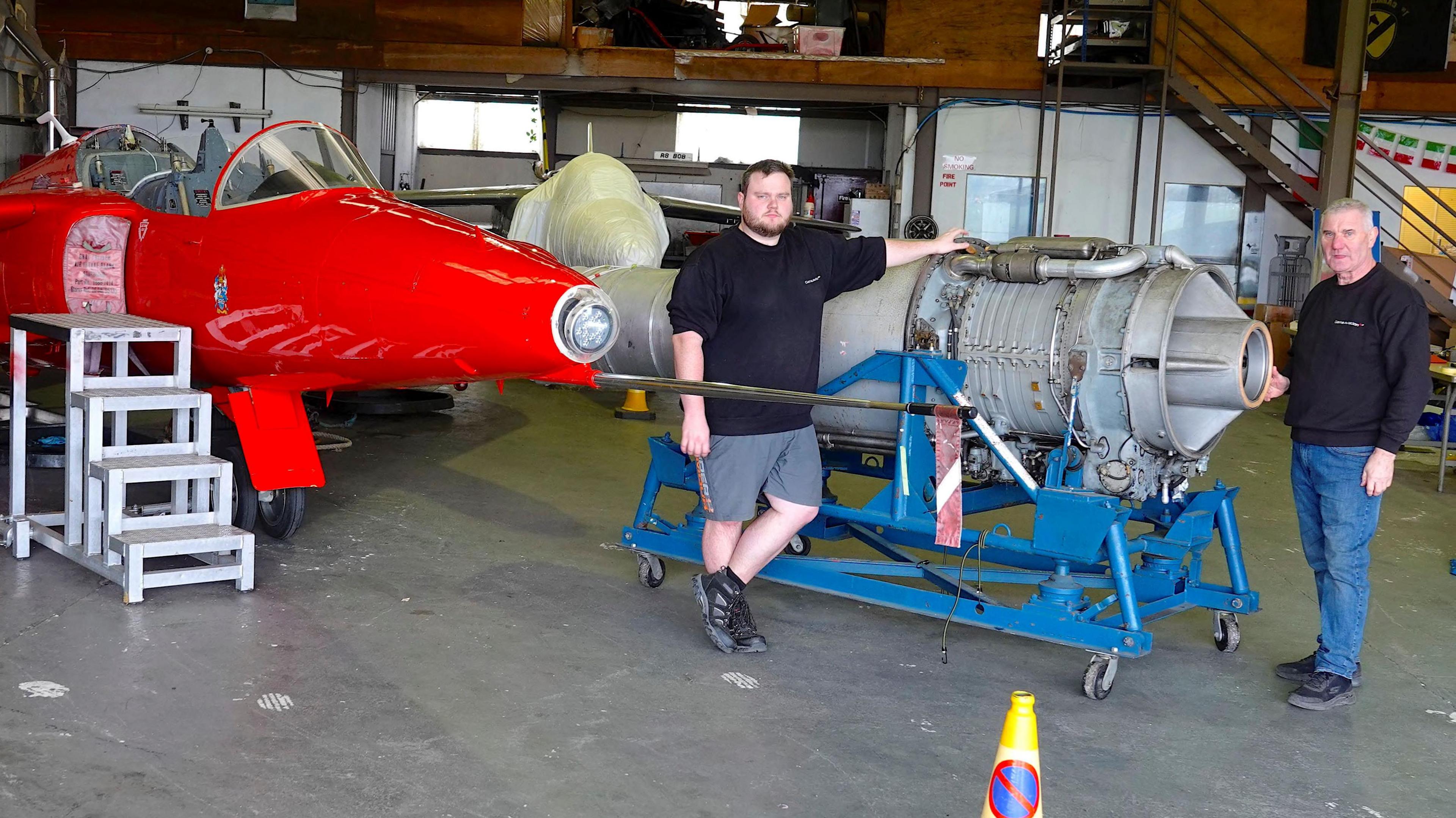 Senior engineer Connar Holland (left) and chief engineer Peter Walker (right) stand beside the mark 101 Orpheus engine. The engine is grey and stands on a blue rig. To the left is a red Gnat aircraft.