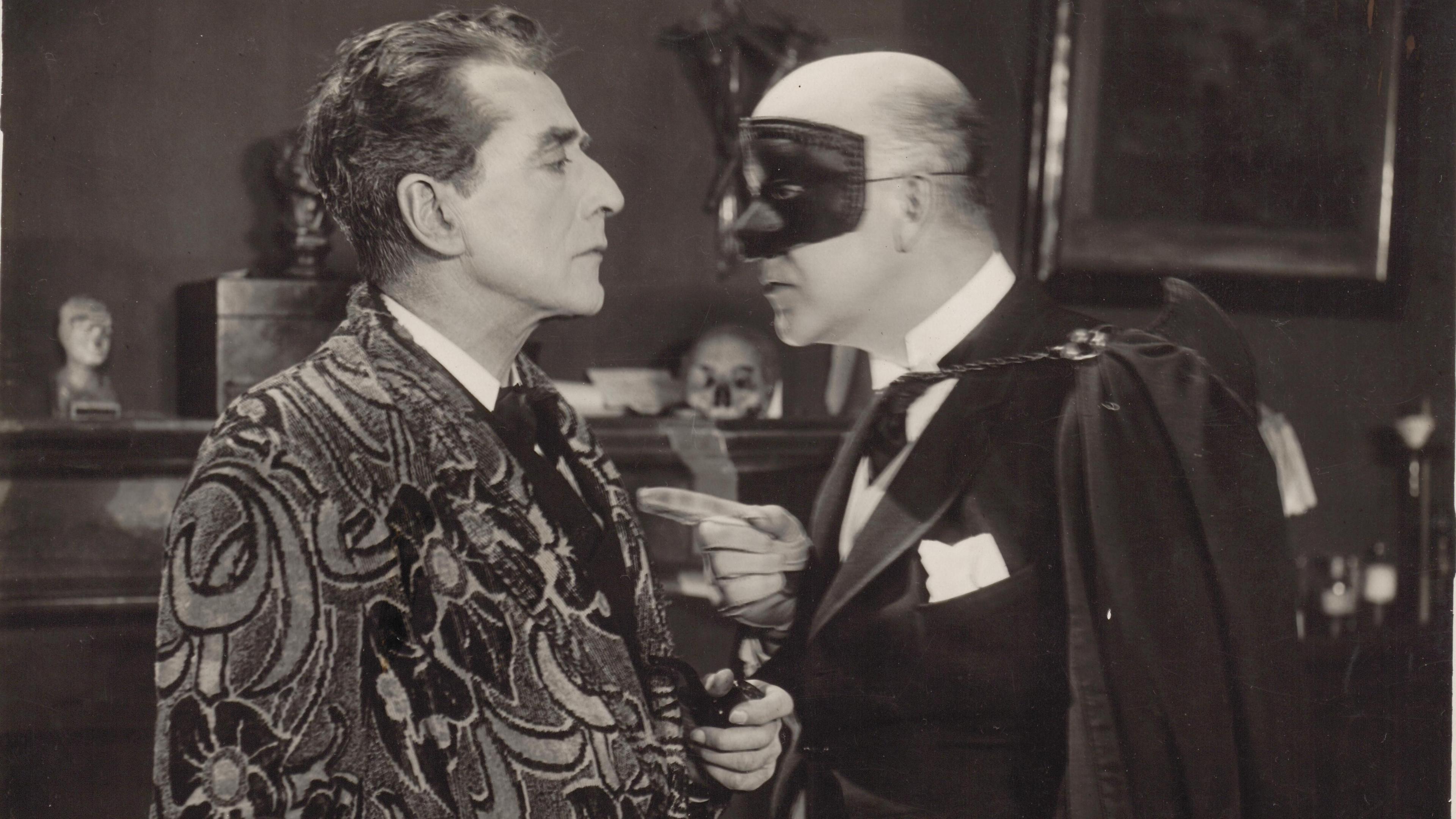 Black and white photograph of a man in an elaborately patterned dressing gown, confronted by a man wearing a cape, gloves and a opera mask 