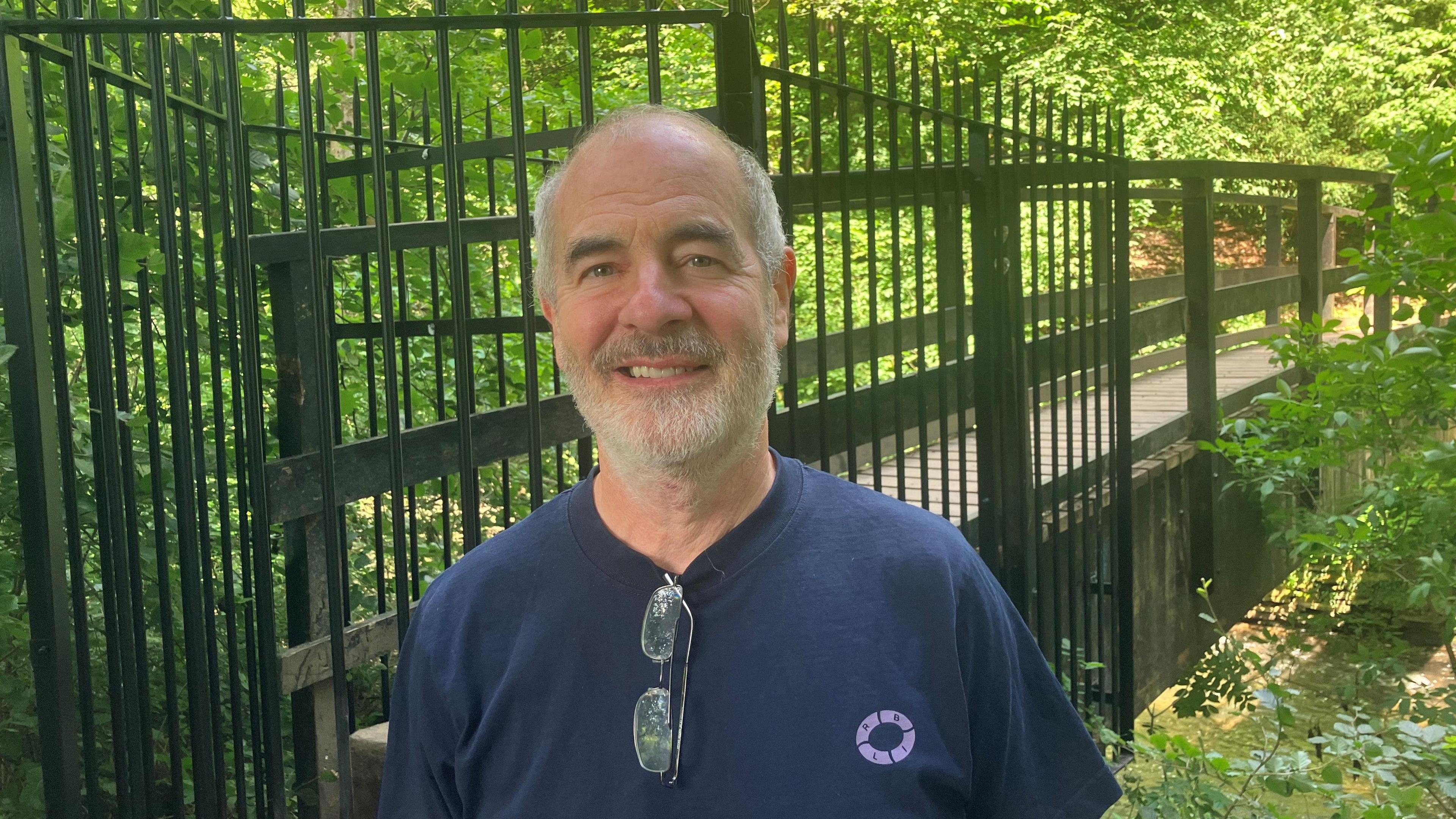 Photograph of Toby Gold, the Chair of Wilmslow Striders running club. With short grey hair and bald on top, he is standing in front of the closed Giant's Castle Bridge in Styal Woods. He's wearing a dark blue t-shirt and has his glasses tucked into the neck of his t-shirt.
