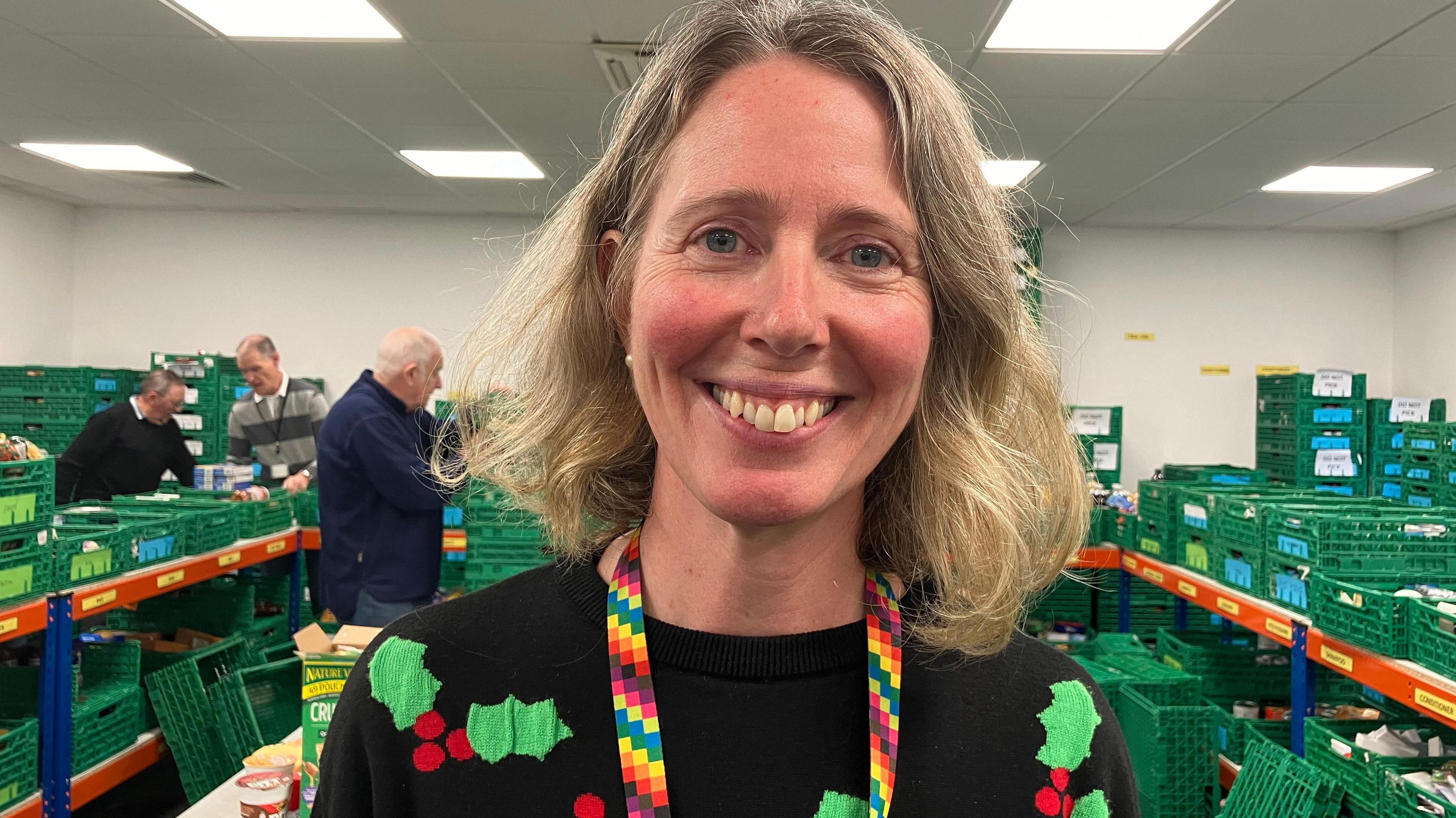 Annette Medhurst is smiling at the camera, with blonde shoulder length hair and blue eyes. She is wearing a black Christmas jumper with green holly and red berries. In the background is a warehouse full of green crates holding food.