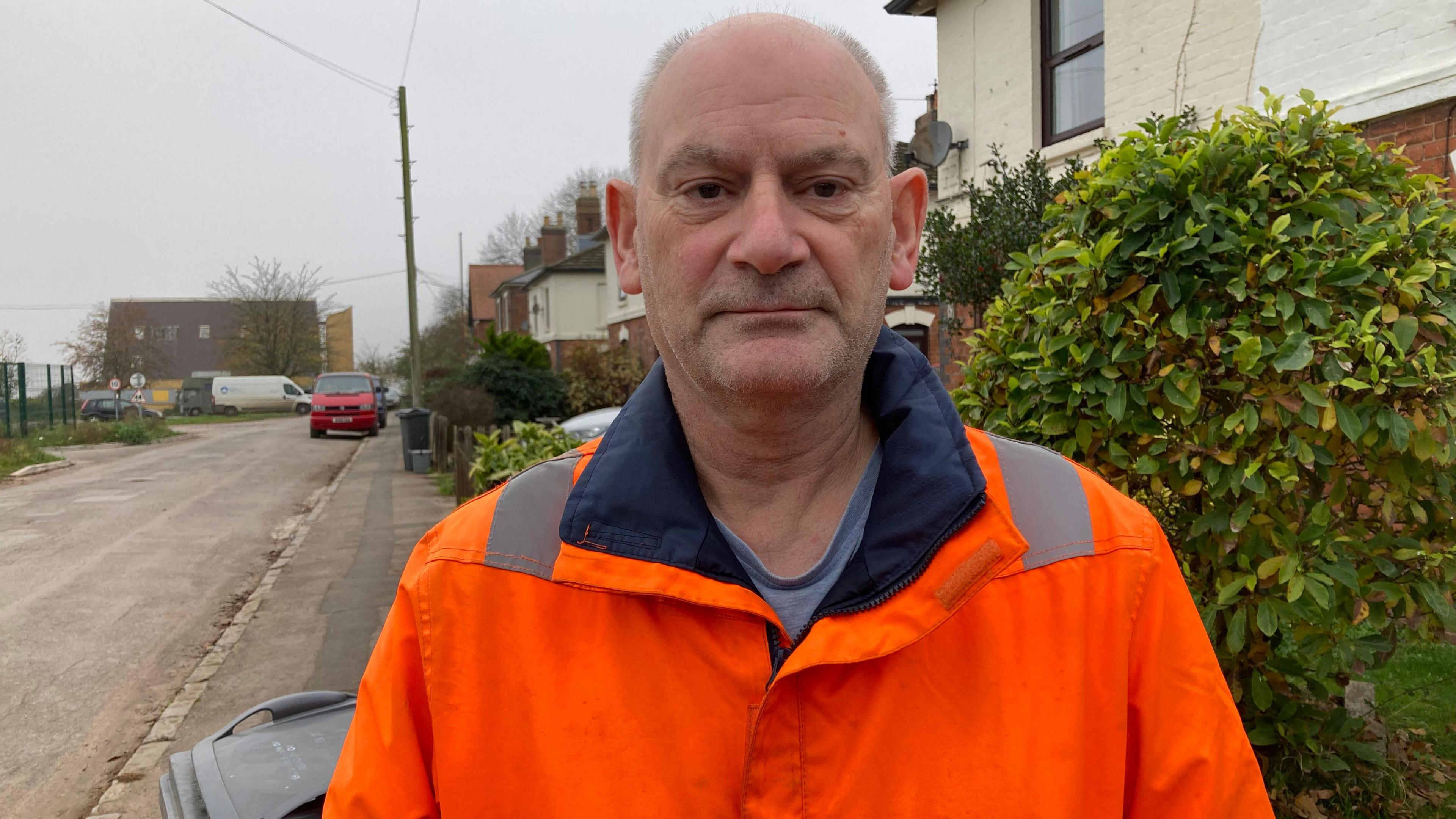 An older man man wearing an orange hi-vis jacket stands on a residential road in an industrial area