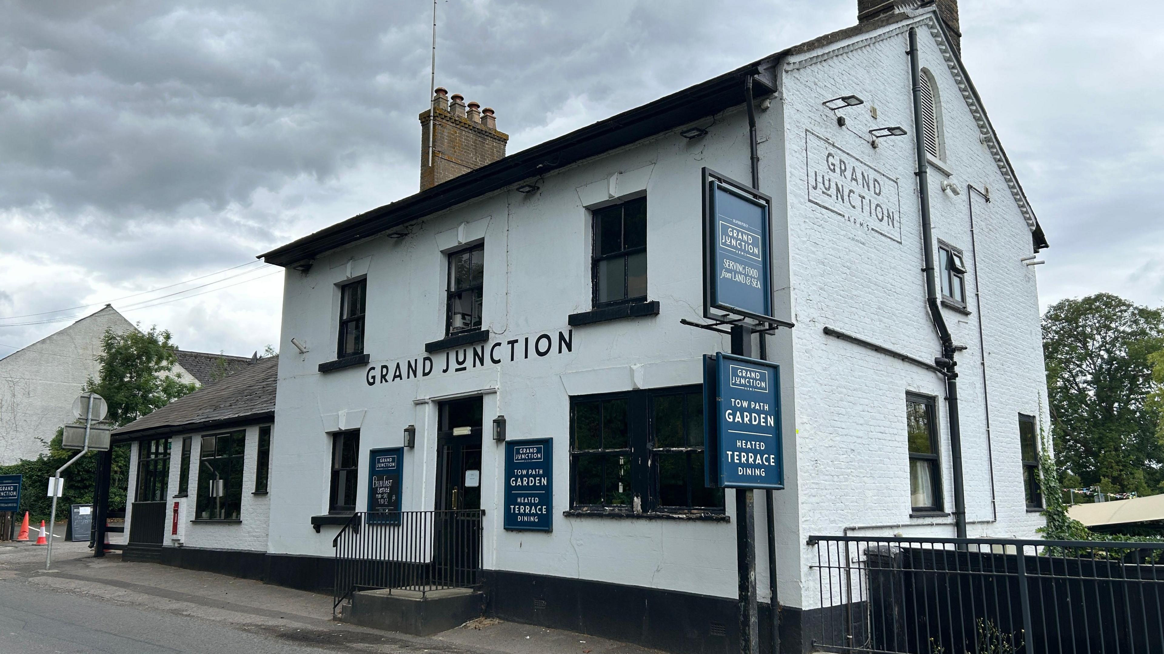 The outside of the Grand Junction, Tring, showing a white pub building, with black window frames, blue signs. There are railings to the right.