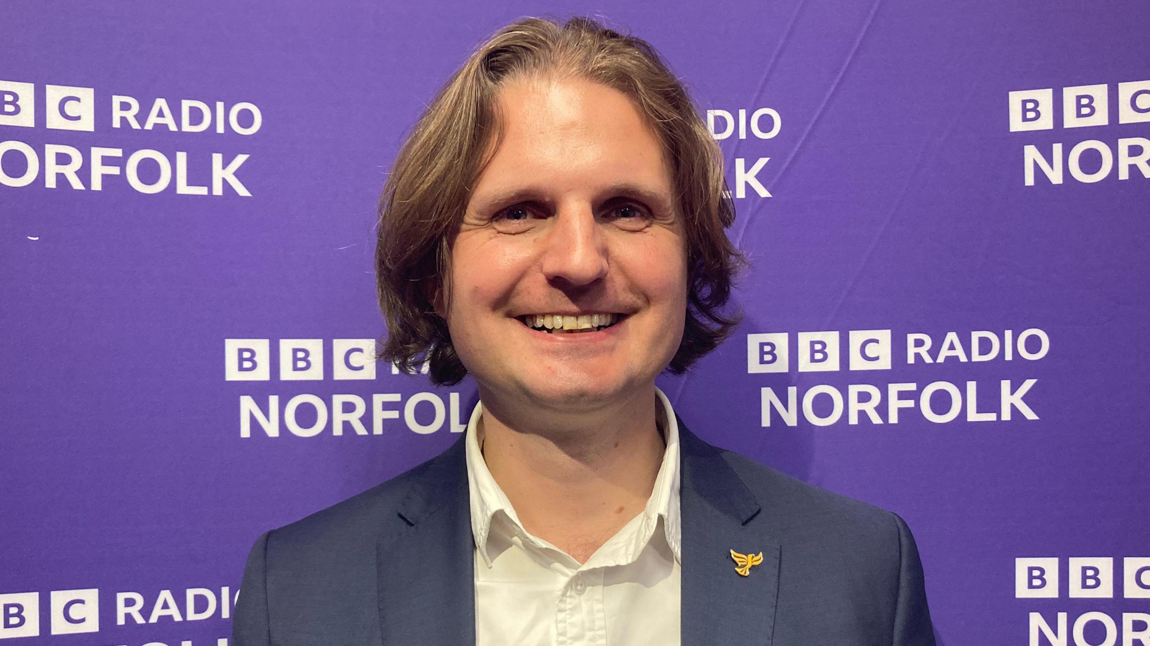 Steffan Aquarone, a man with brown hair who is standing inside a BBC Radio Norfolk radio studio. He is wearing a white shirt and a navy suit jacket and is looking directly at the camera and smiling.