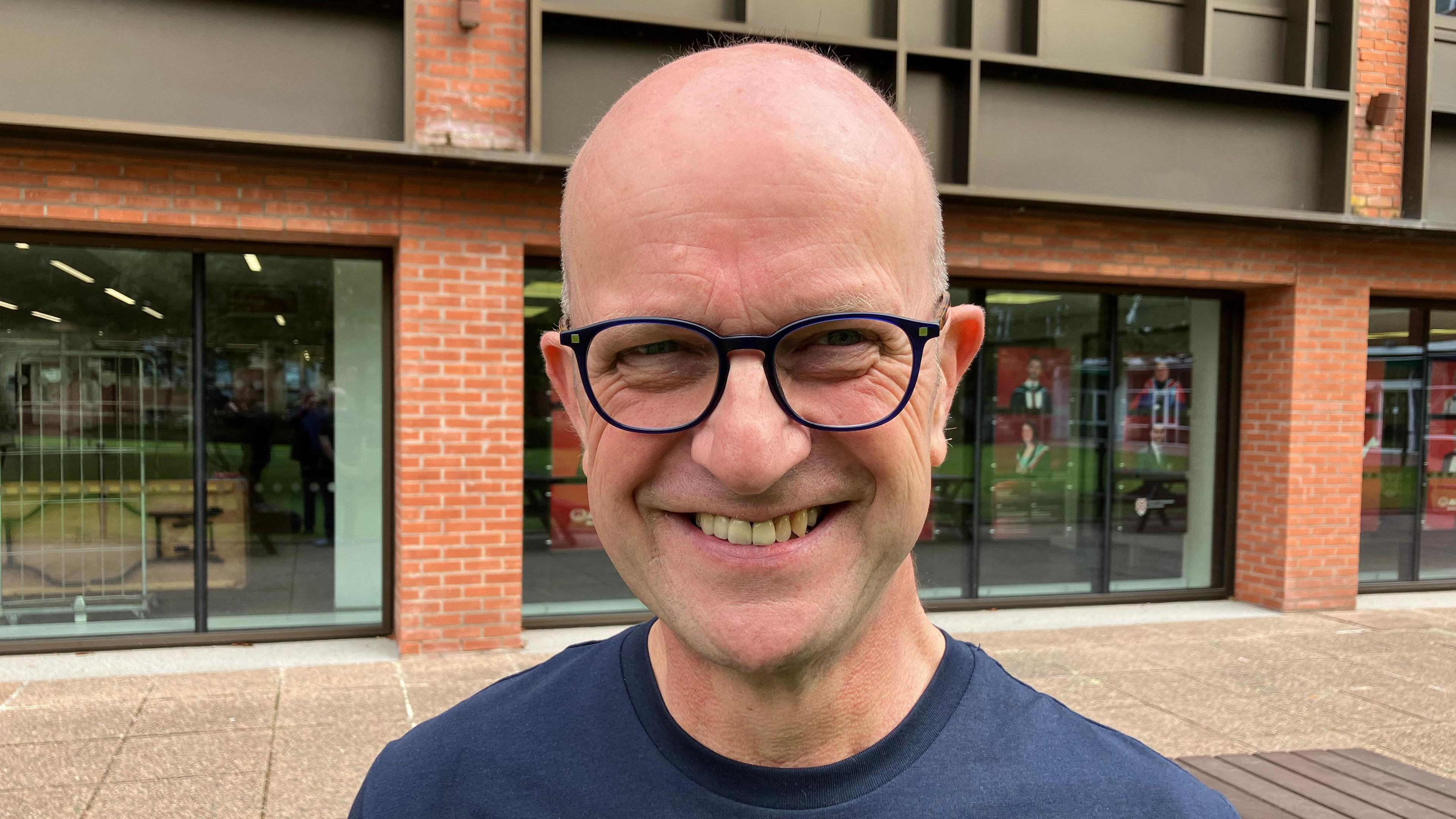 Paul is standing in front of three windows and a red brick building.

He is bald and is wearing black rimmed glasses. 

He is smiling at the camera and is wearing a blue turtle neck t-shirt. 