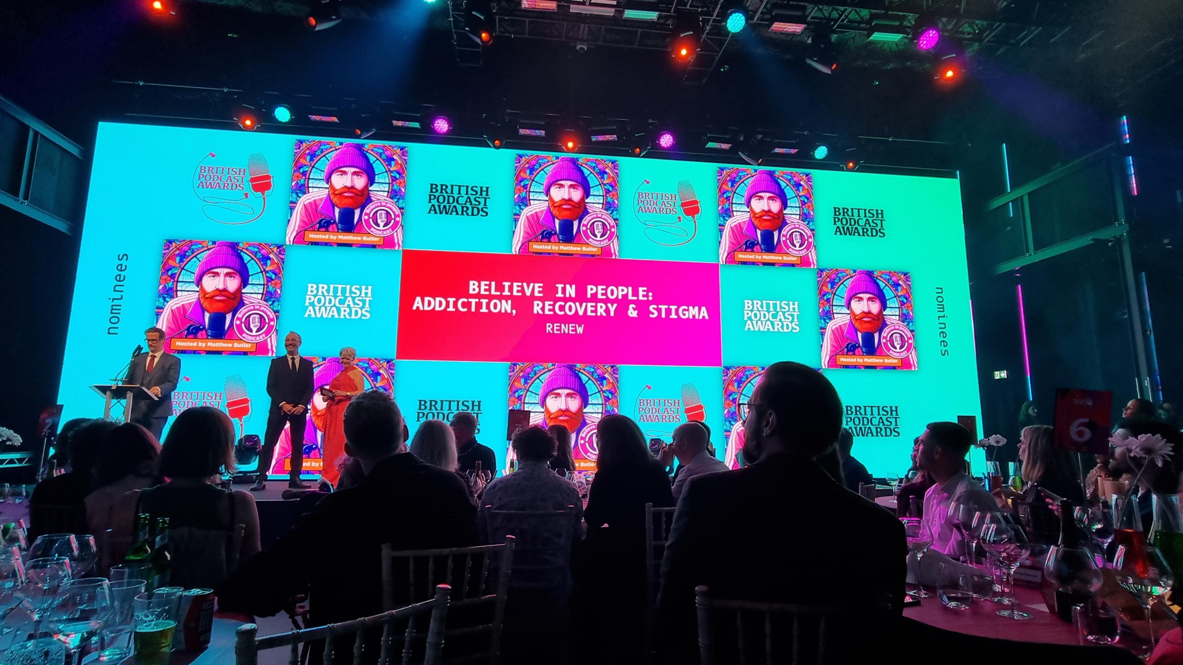 A big screen showing the thumbnail for the podcast Believe in People: Addiction, Recovery & Stigma. Multiple people are sat around tables watching three people on stage announcing an award. 