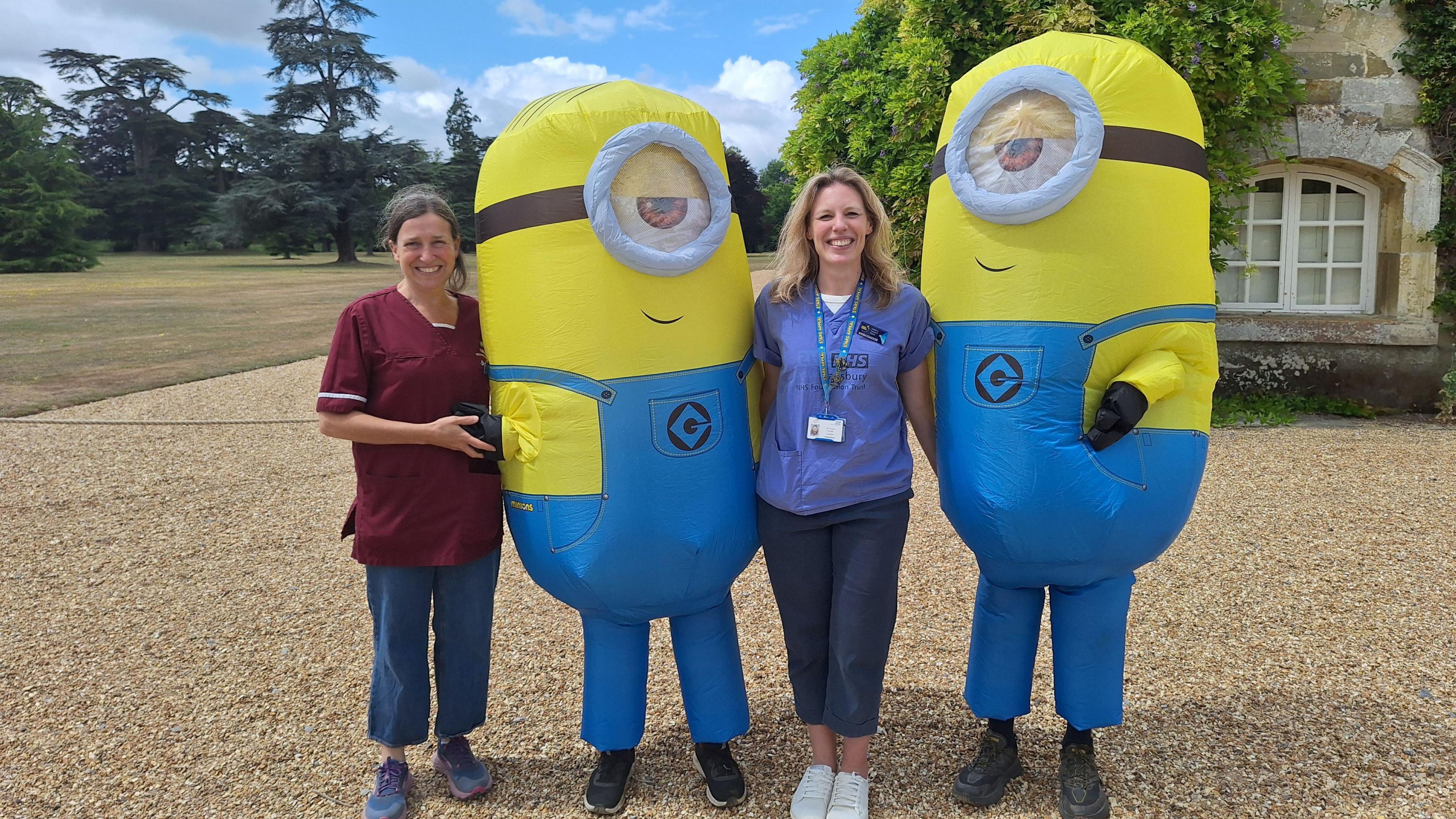 Four people standing together outside a country house. Two are wearing inflatable Minion costumes, with blue dungarees on the yellow, one-eyed characters.