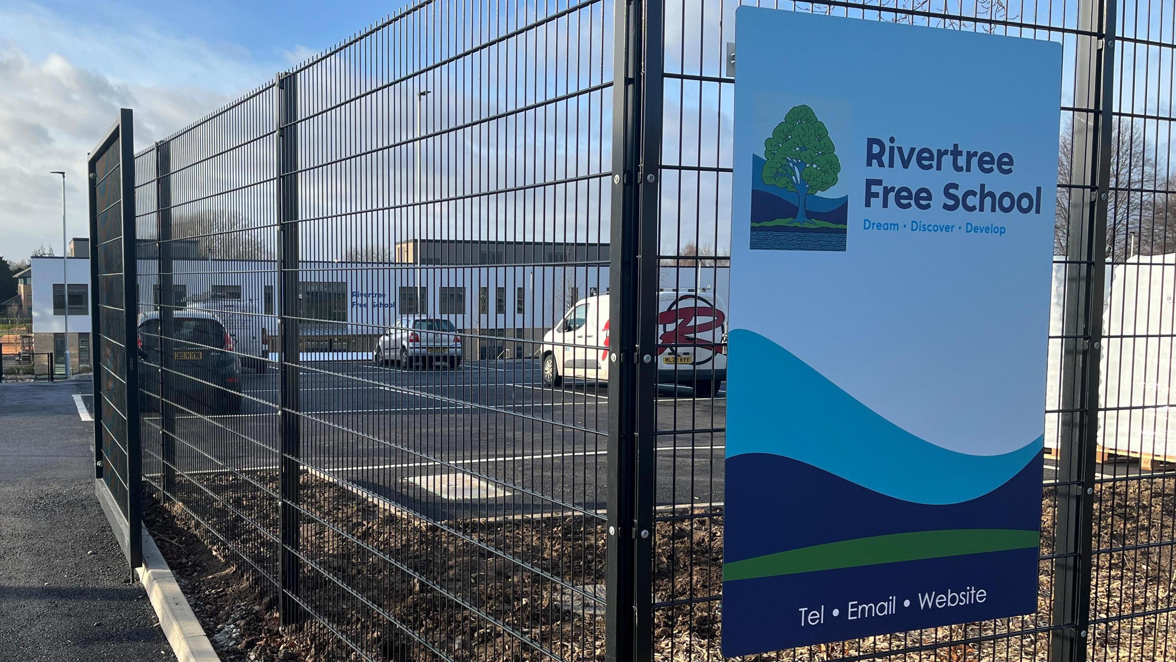 The outside gates of a school with a sign saying "Rivertree Free School" with a car park in the background and the school building itself beyond the car park.