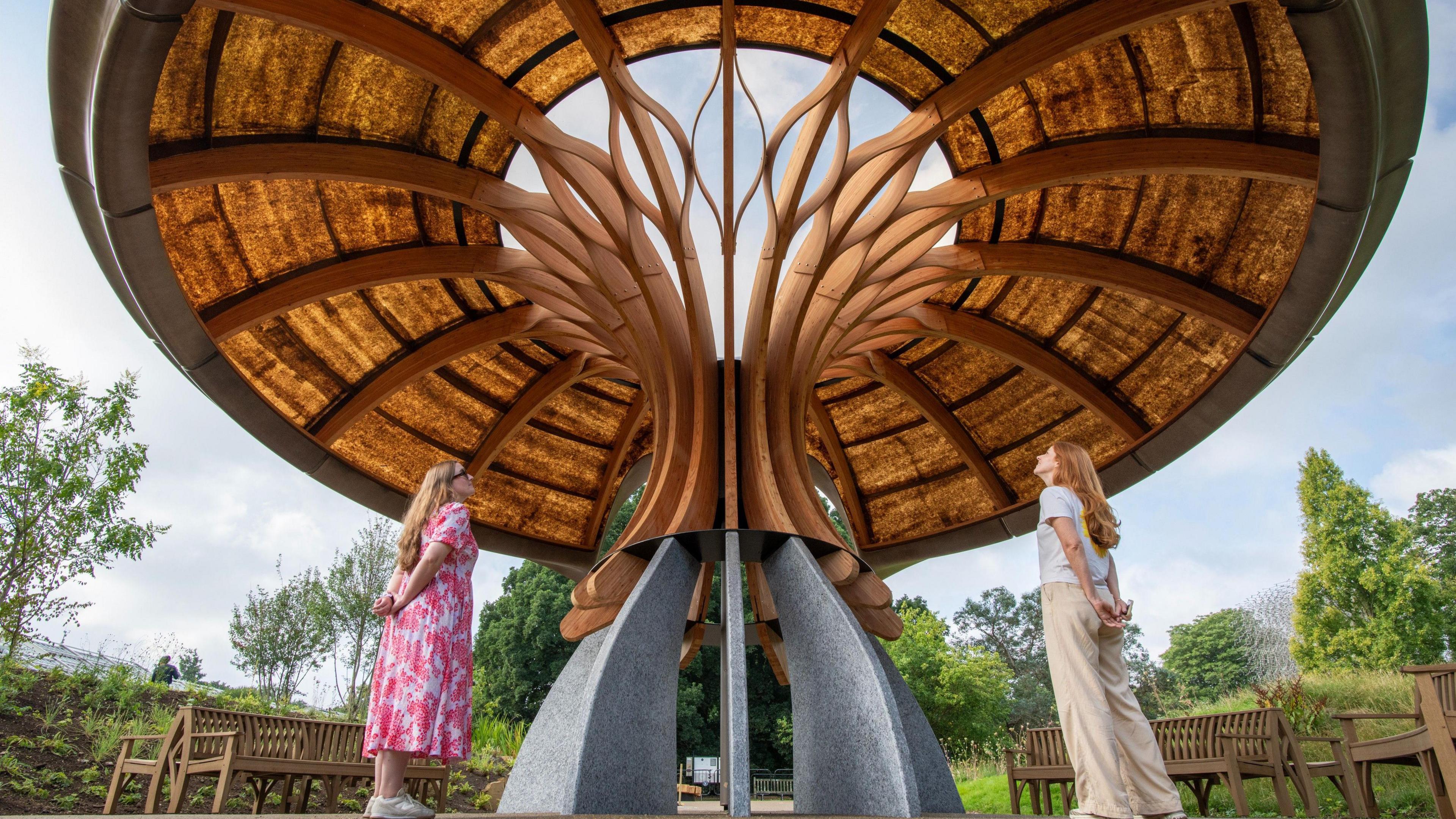 Two women with long hair standing on either side of a large, artistic wooden structure with a curved roof and wing-like designs. There are trees in the background. The photo is taken from the ground up.