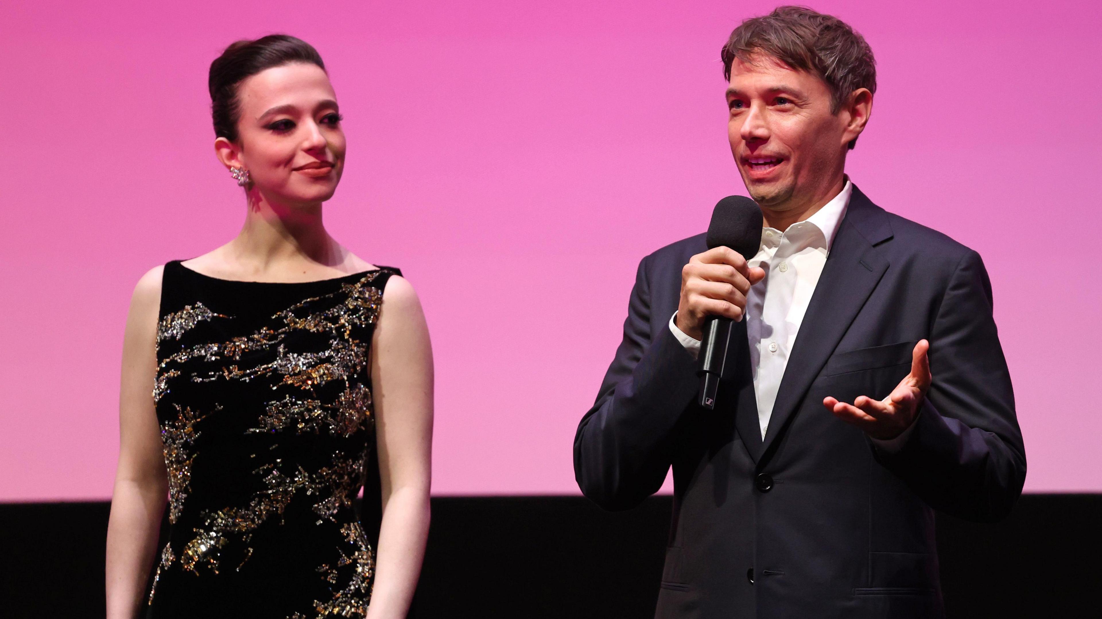 Mikey Madison and Director Sean Baker on stage during the "Anora" Headline Gala during the 68th BFI London Film Festival at The Royal Festival Hall on October 11, 2024 in London, England. They are onstage introducing the film at the premiere. Baker is holding a microphone and talking to the audience. 