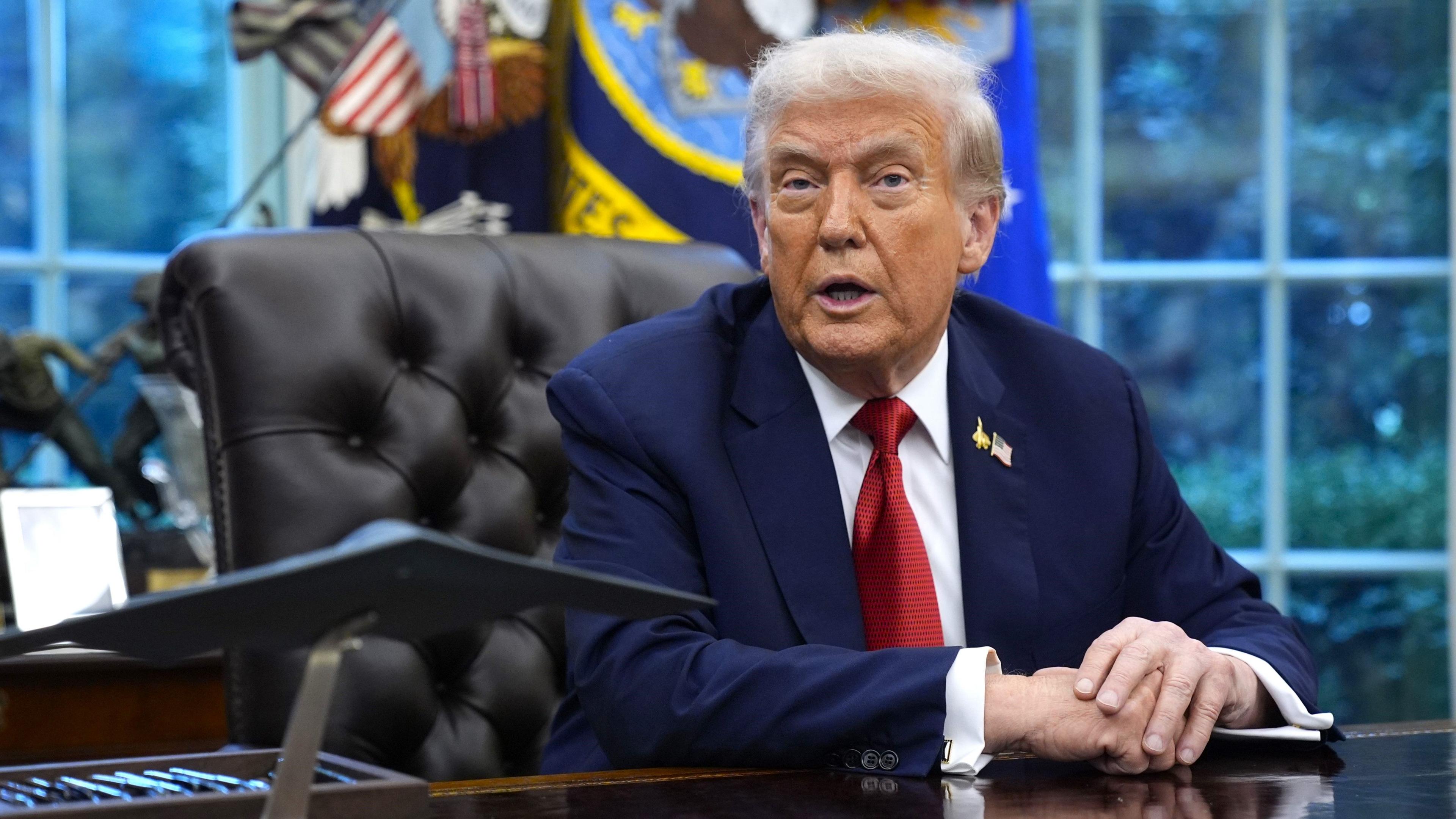 US President Donald Trump sits at his desk in the Oval Office. He wears a navy suit and red tie and rests his hands on the desk.