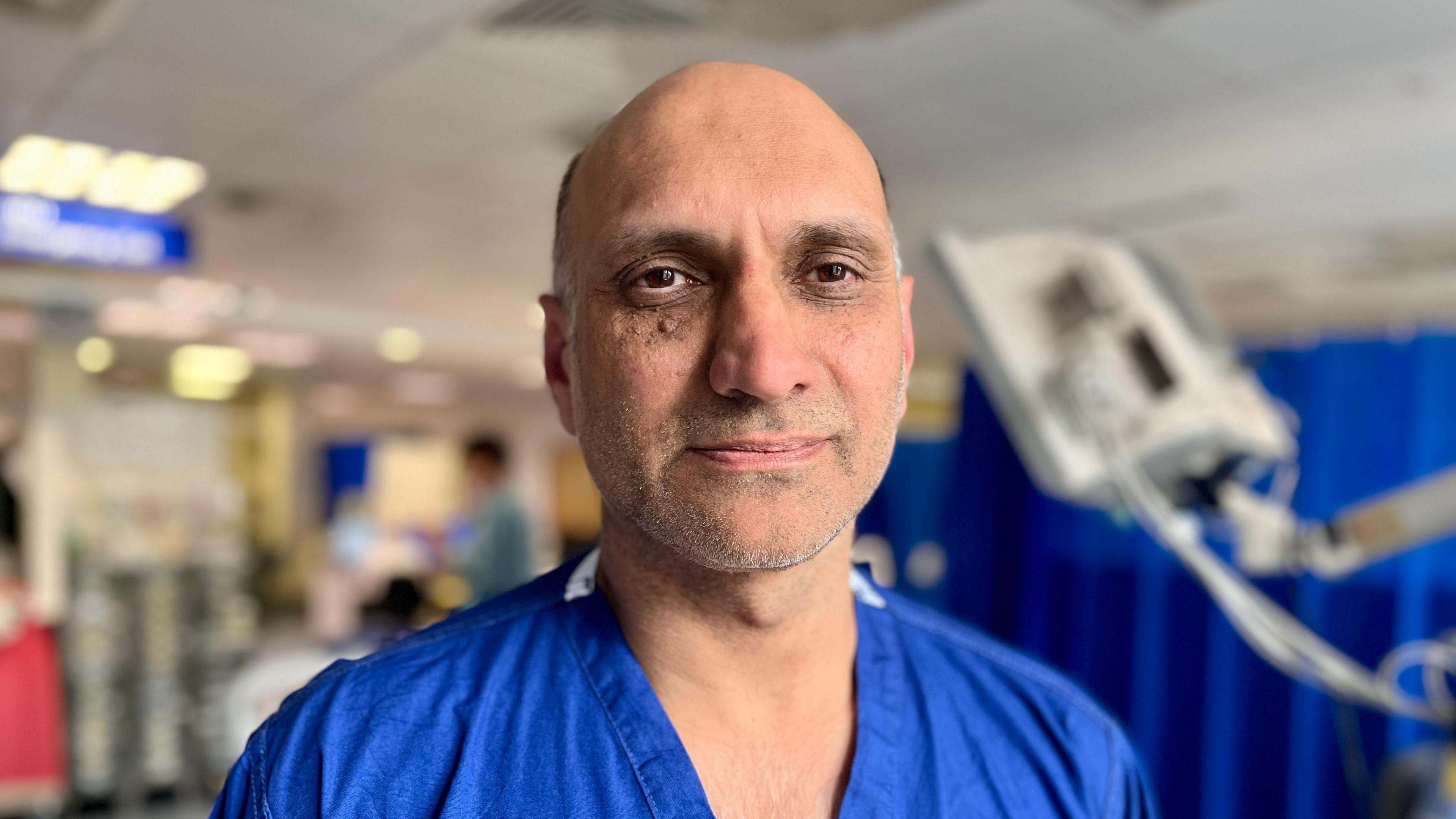 Dr Parvez Moondi stands in front of the critical care department at the Norfolk and Norwich Hospital. He is wearing blue scrubs with no PPE. He has a balding head and you can see his head and shoulders. He is looking straight at the camera and has brown eyes.