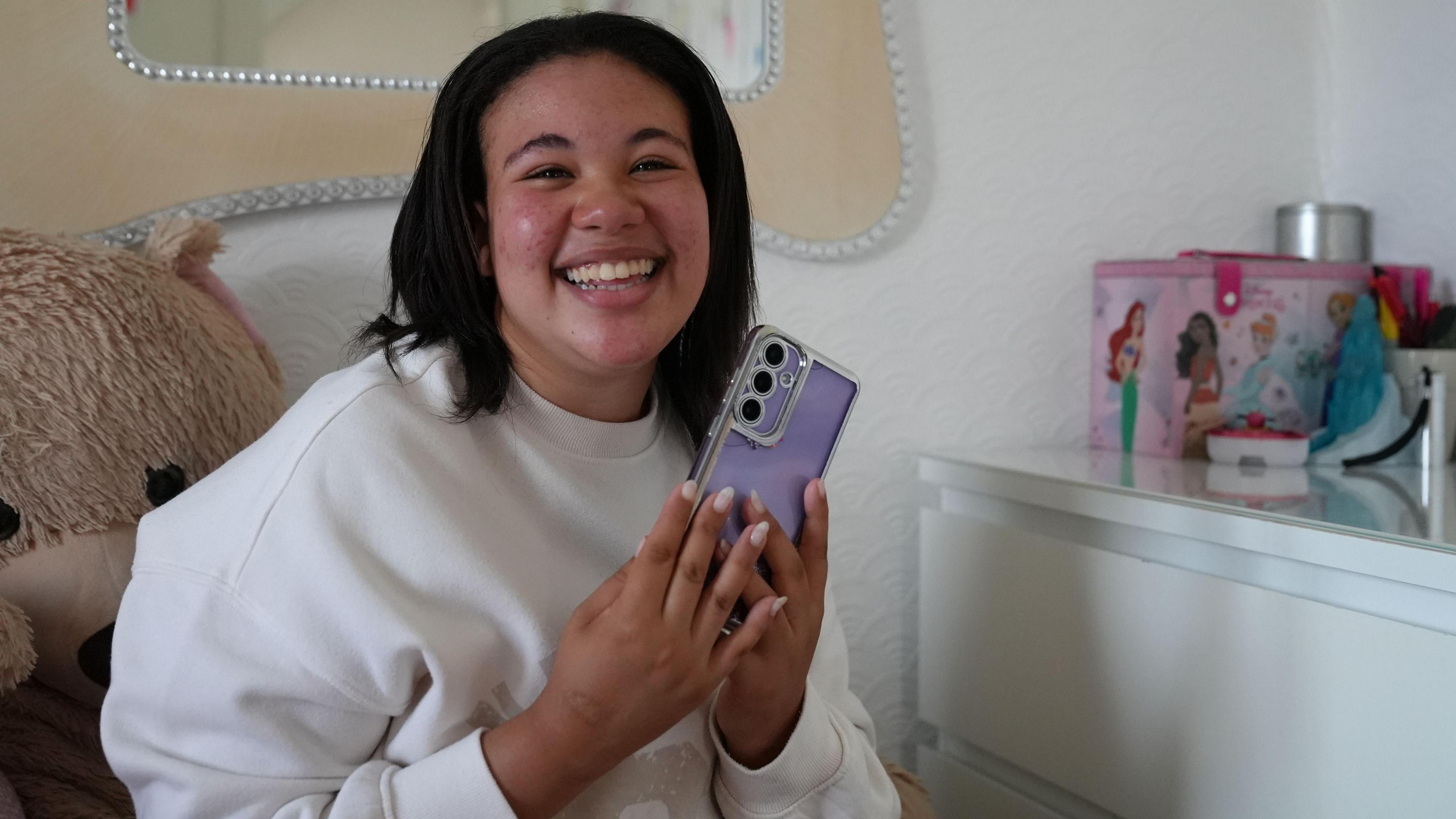 A teenage girl clutches her phone which has a purple phone case and smiles while sitting on her bed in her bedroom.