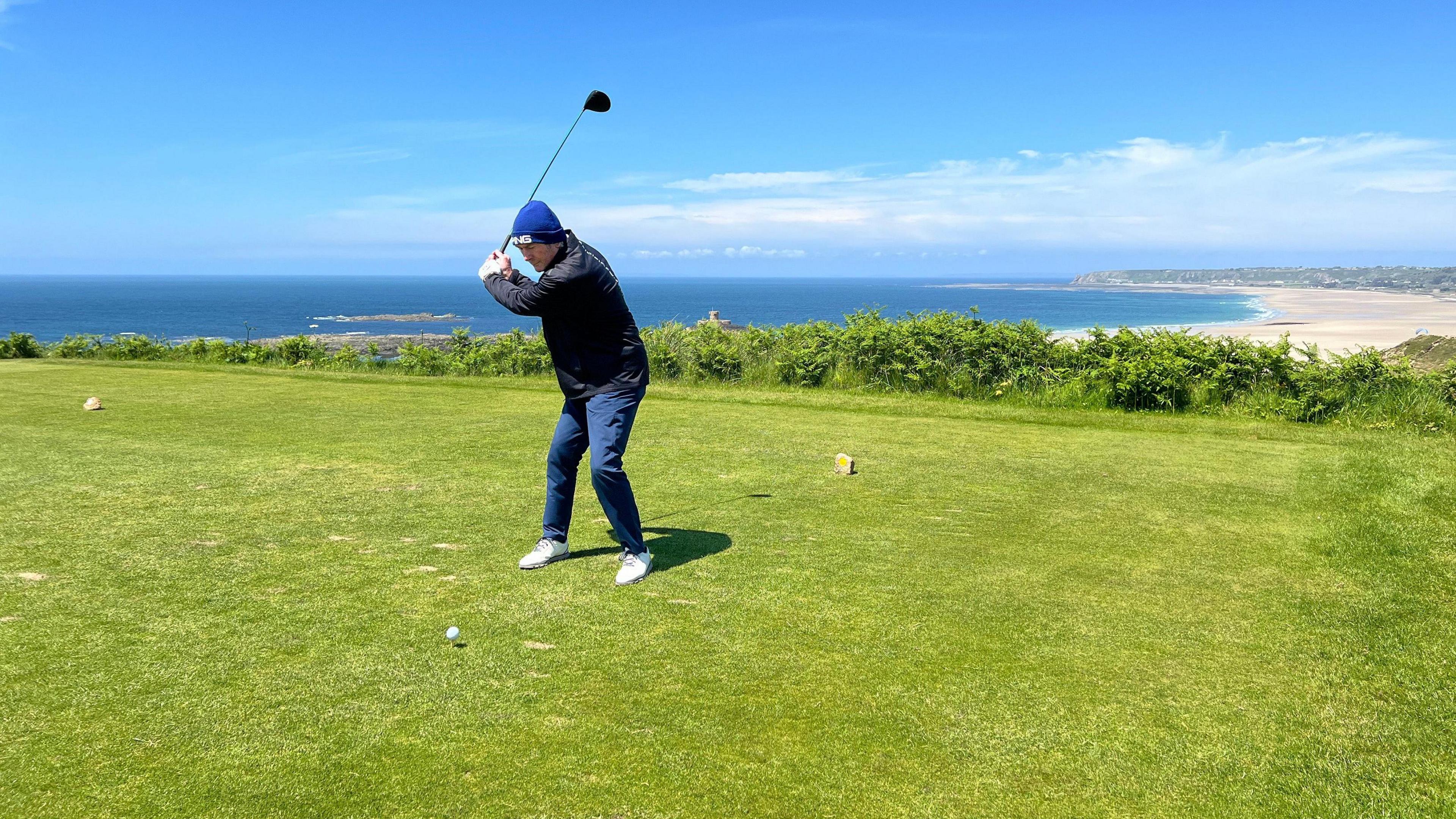 Pat raises his golf club above his head to tee off and behind him a long beach extends into the background