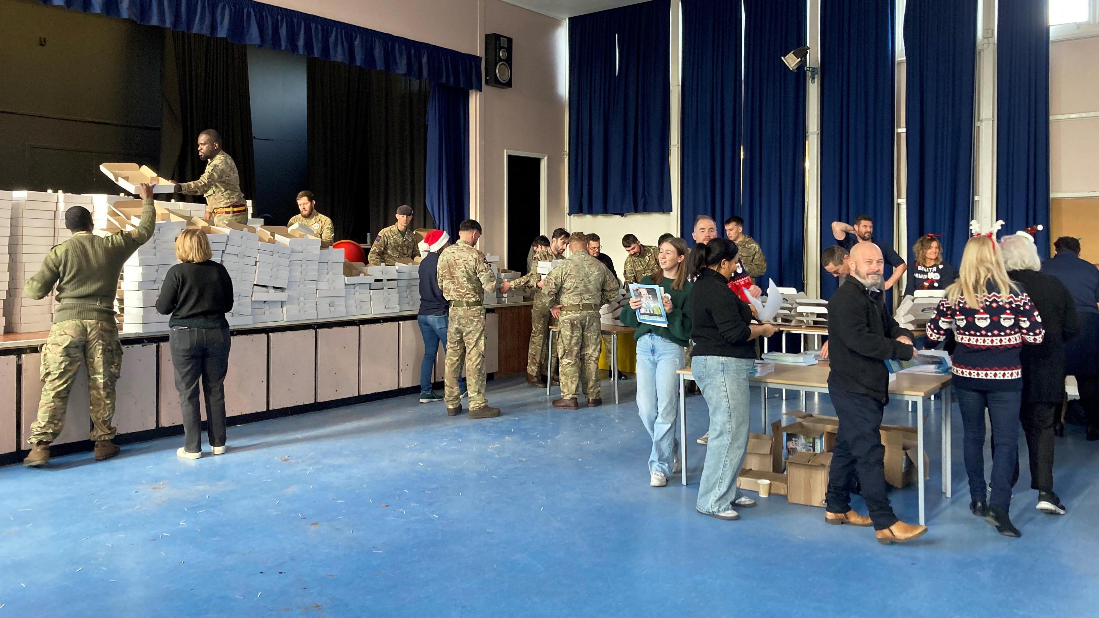 Army personnel and volunteers in a hall in a line system putting together the boxes.