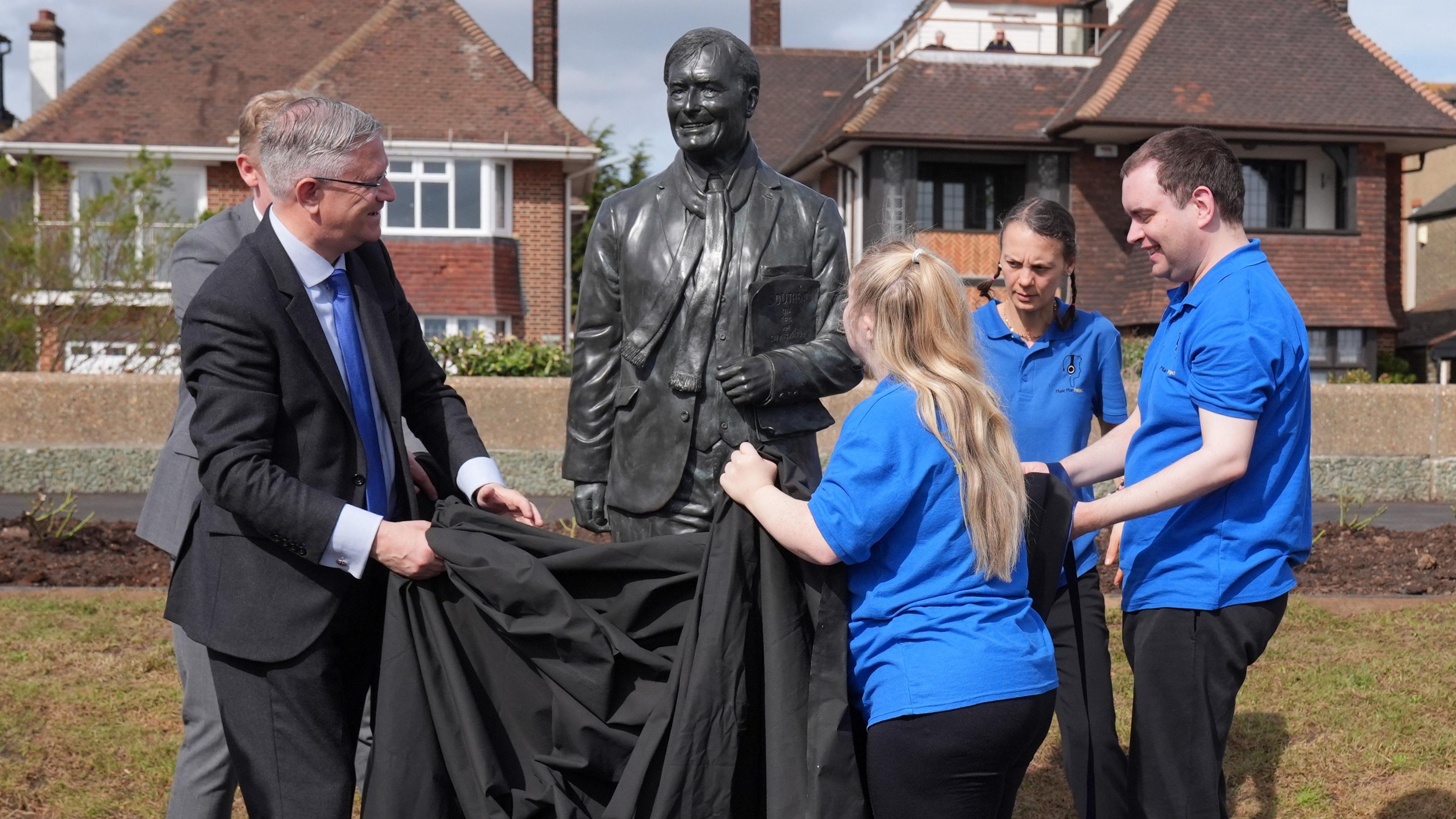 Three people wearing blue polo shirts and a man wearing a black suit pulling a black sheet off of a statue of Sir David Amess on Chalkwell seafront. They are joined by a man in a grey suit. Two houses are in the background.