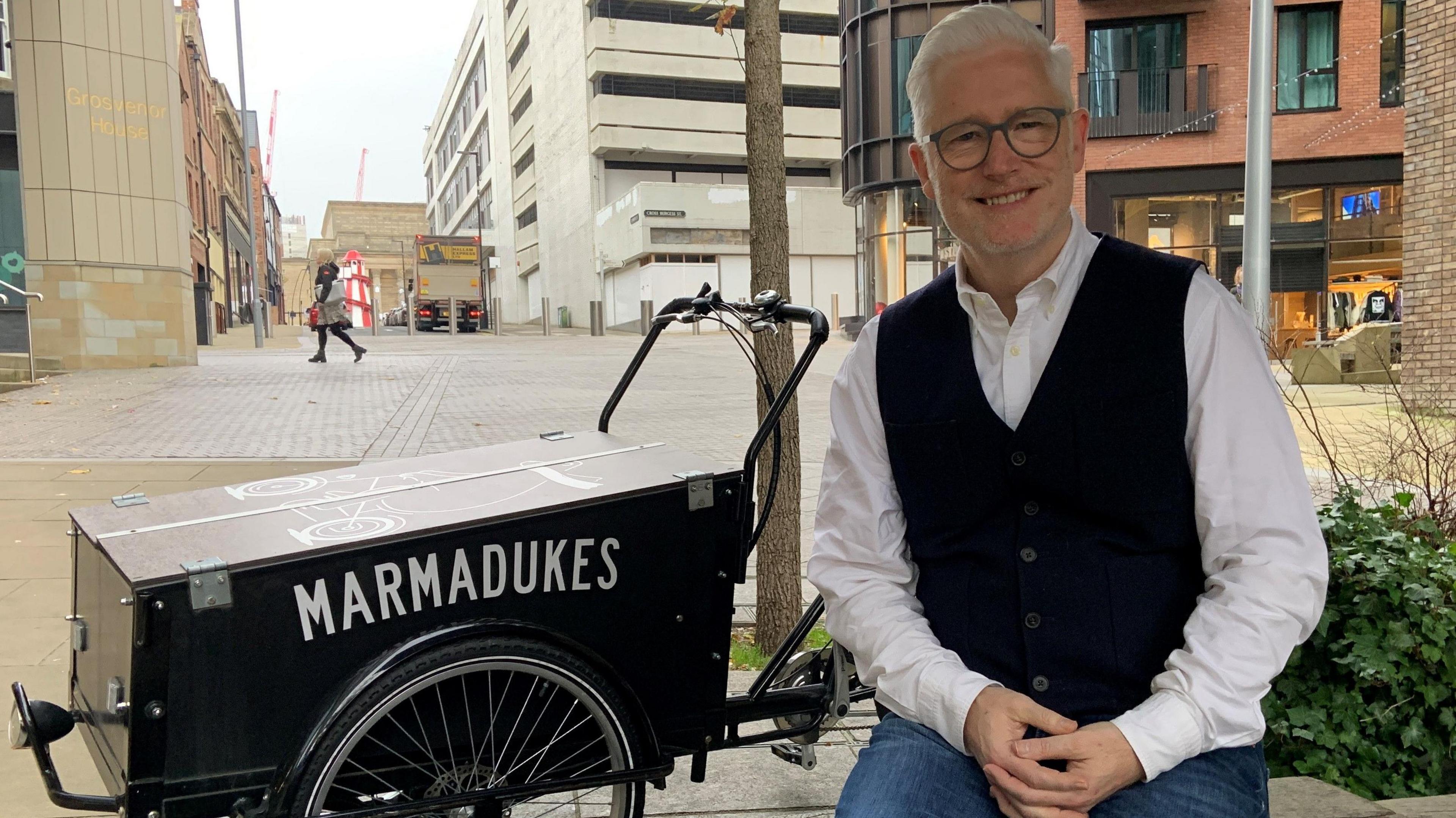 A man with white hair and glasses is smiling and wearing a white shirt and black waistcoat. He is sitting outdoors in the city centre. Next to him is an old fashioned small black wagon with Marmadukes written on the side in white letters