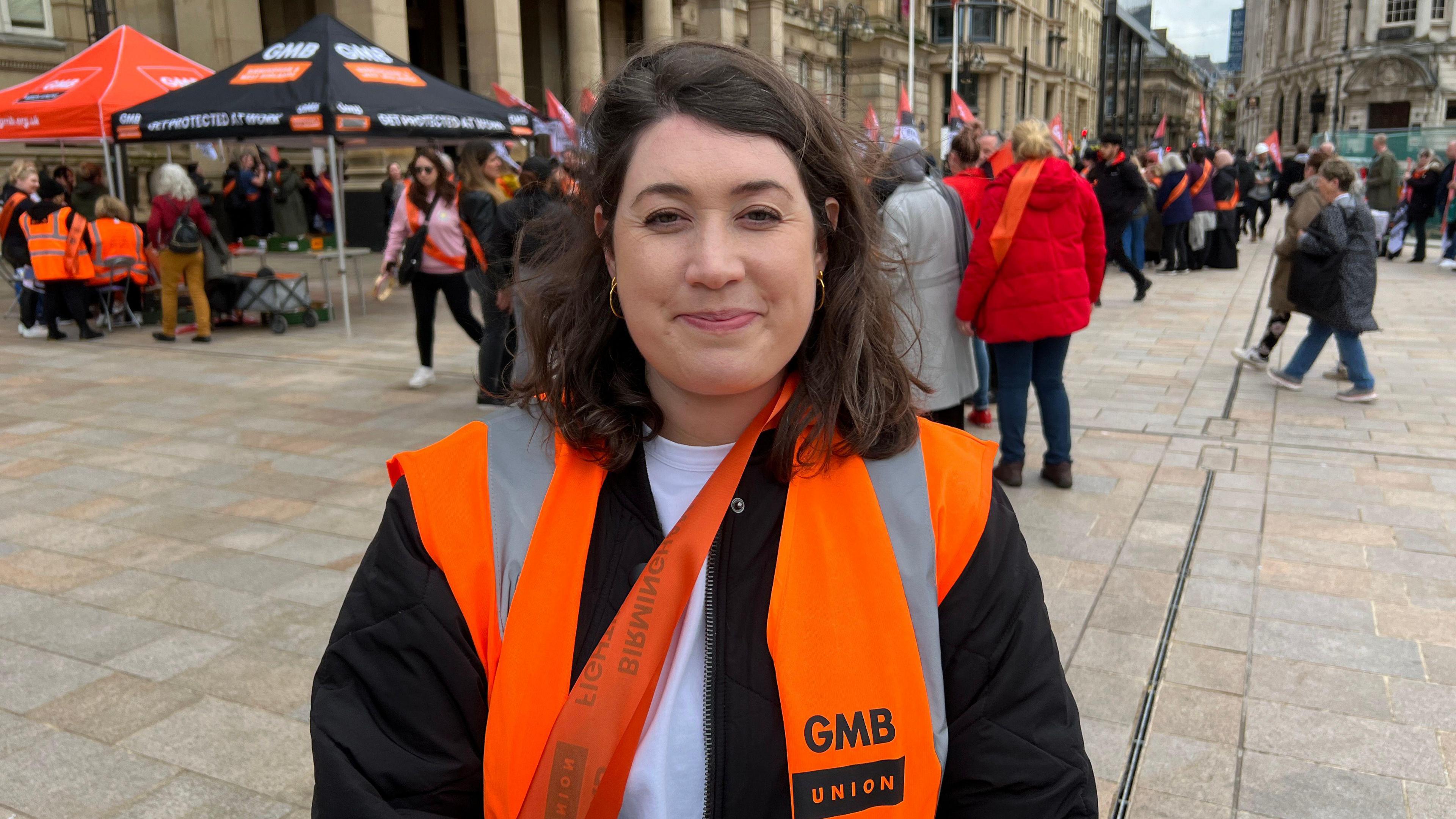 A smiling woman with dark hair to her shoulders stands in a paved square in front of a crowd of people. Behind her is a pagoda stand with the GMB Union logo on it