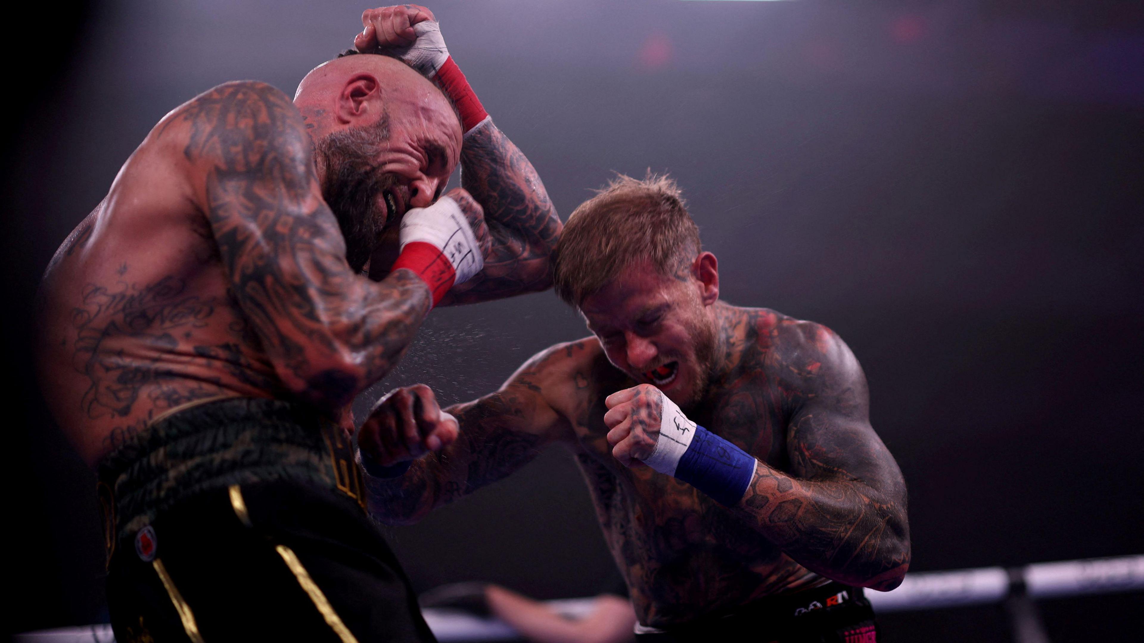 Two men, both with lots of tattoos and one with a shaved head and beard and the other with short ginger hair and no beard, grimace as they fight during a bareknuckle boxing event in Bristol.
