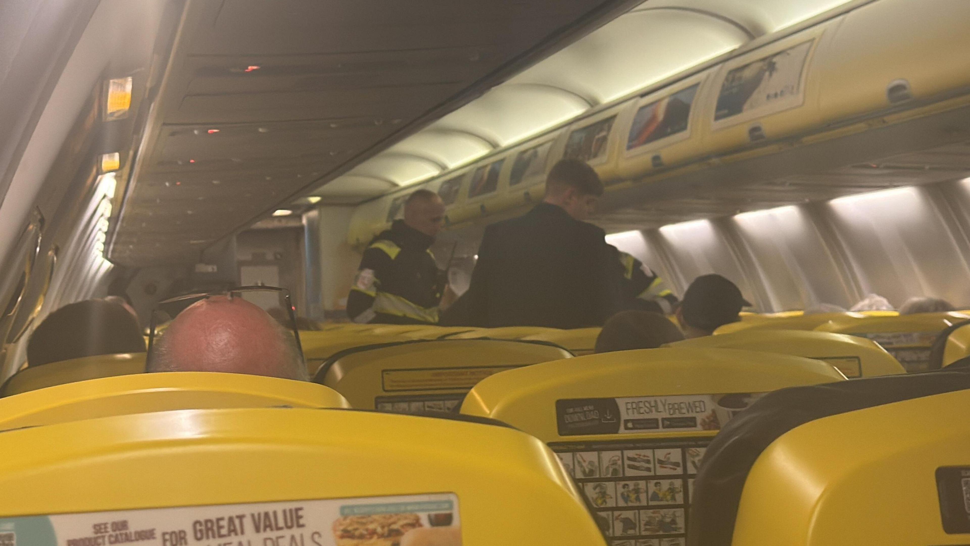 Passenger's view of emergency staff standing at the front of the aircraft cabin. The plane is full and the backs of people's heads can be seen poking up between the rows of bright yellow seats.