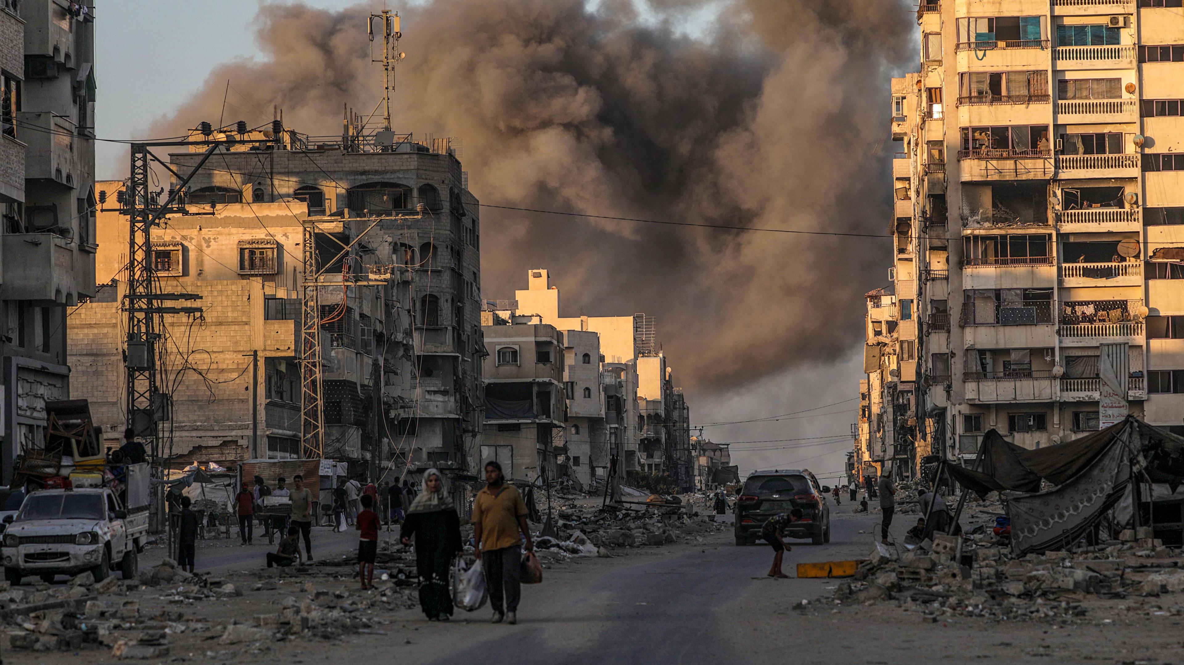 Smoke can be seen rising behind high rise buildings in Gaza. People can be seen walking in the street as well as a couple of cars. There is lots of debris strewn across the ground.