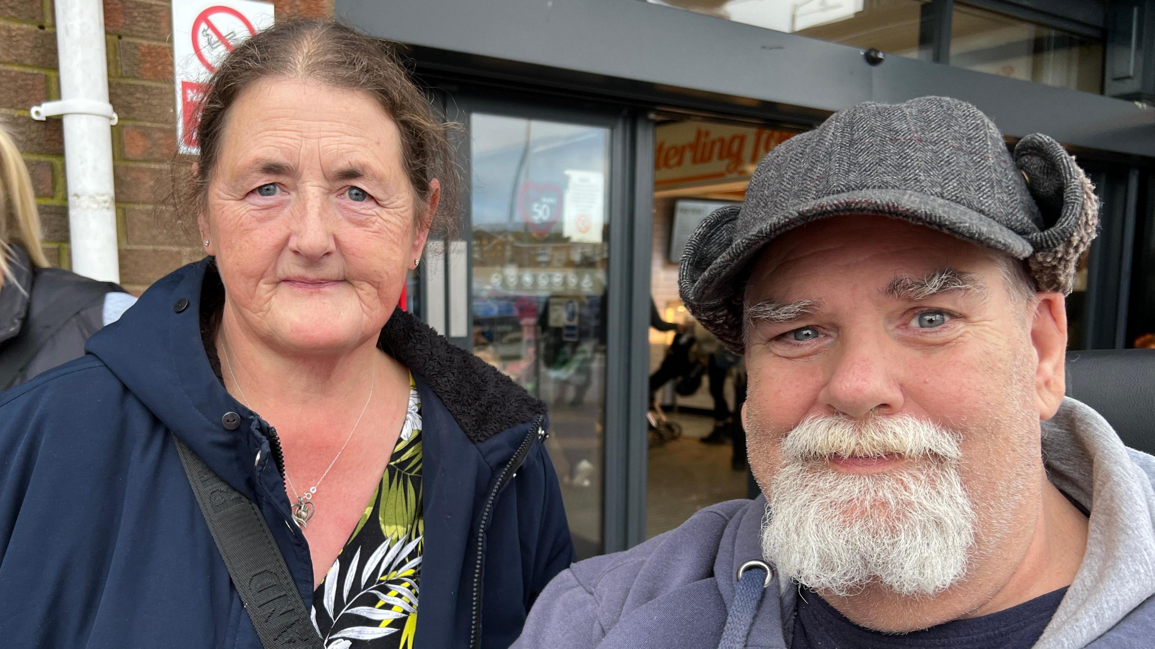 A man sitting on a mobility scooter and a woman standing up pose close together for a photograph. The man wears a grey deer-stalker hat and a blue hooded top and has a white beard. The woman has brown hair pulled back into a ponytail and wears a navy zip-up jacket.