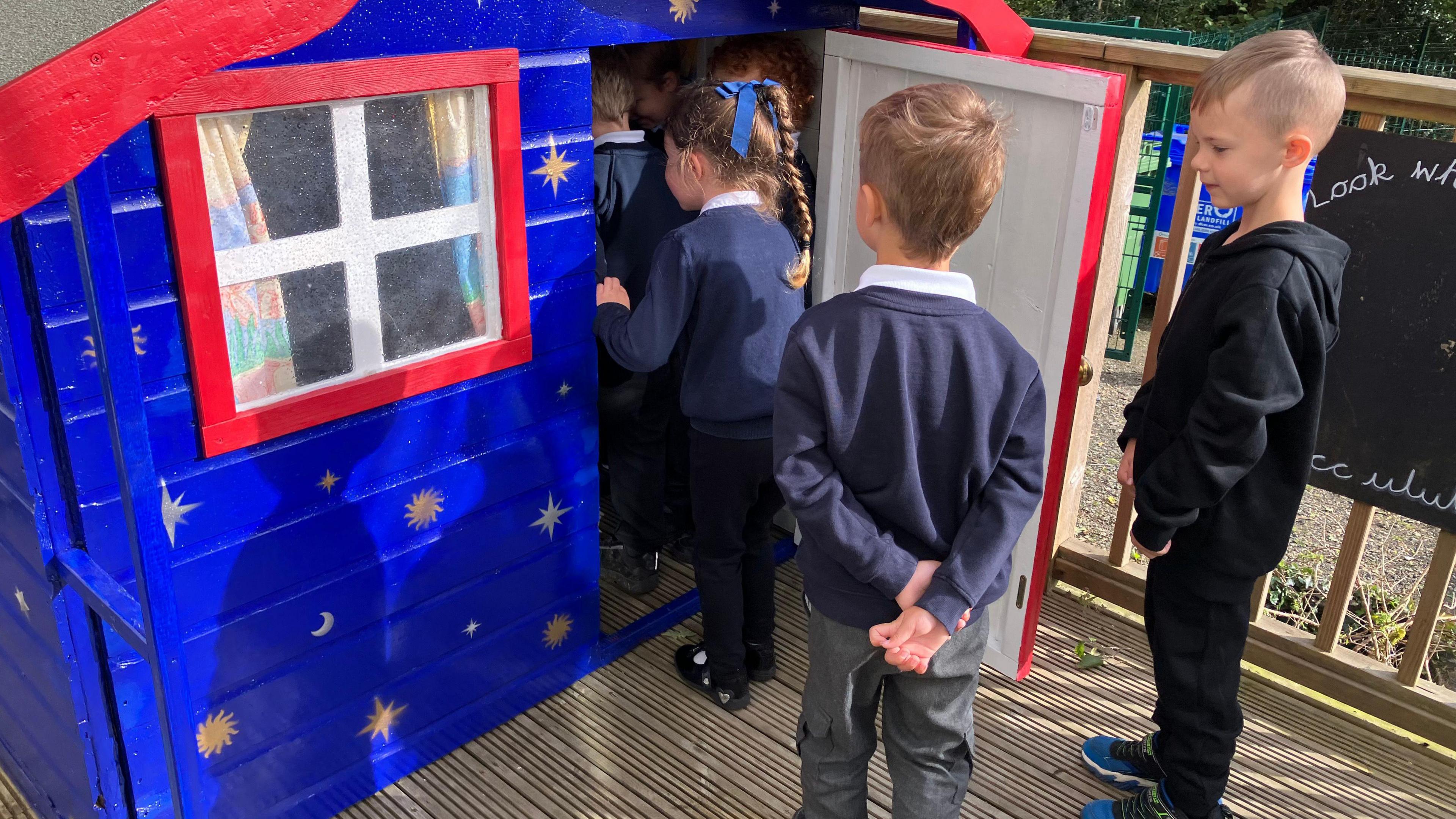 A few primary school children in blue uniforms in front on a blue and red Wendy house waiting to get in, one boy hangs back.
