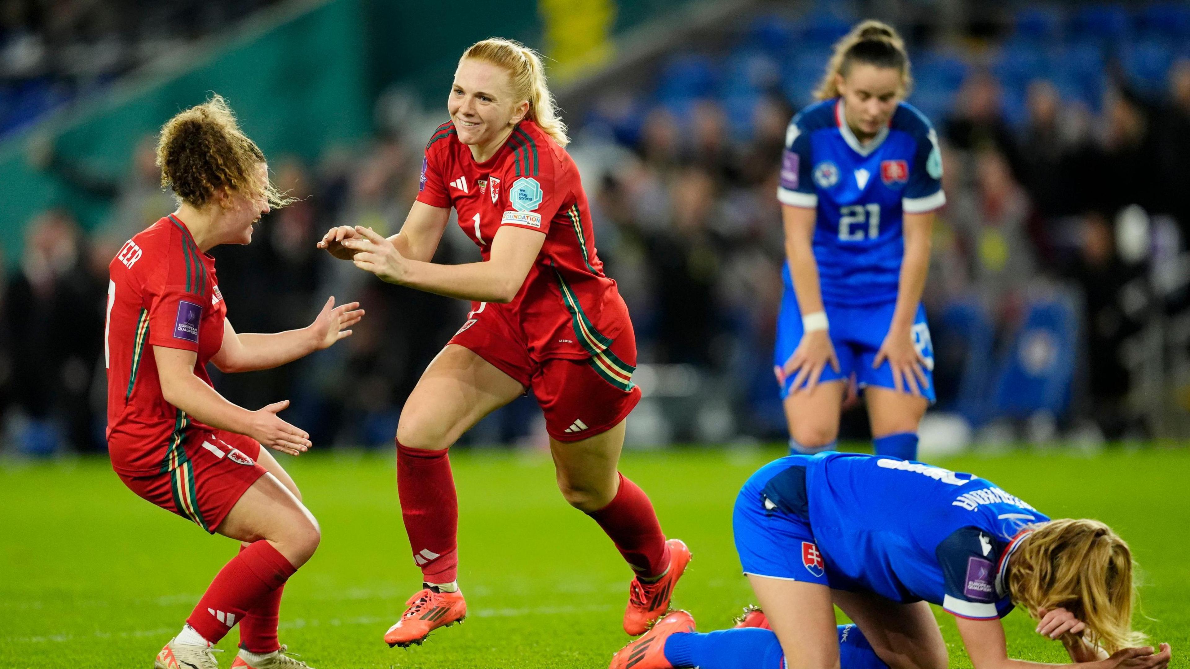 Two Welsh football players run to shake hands after scoring a goal, with two Slovakian players looking disappointed 