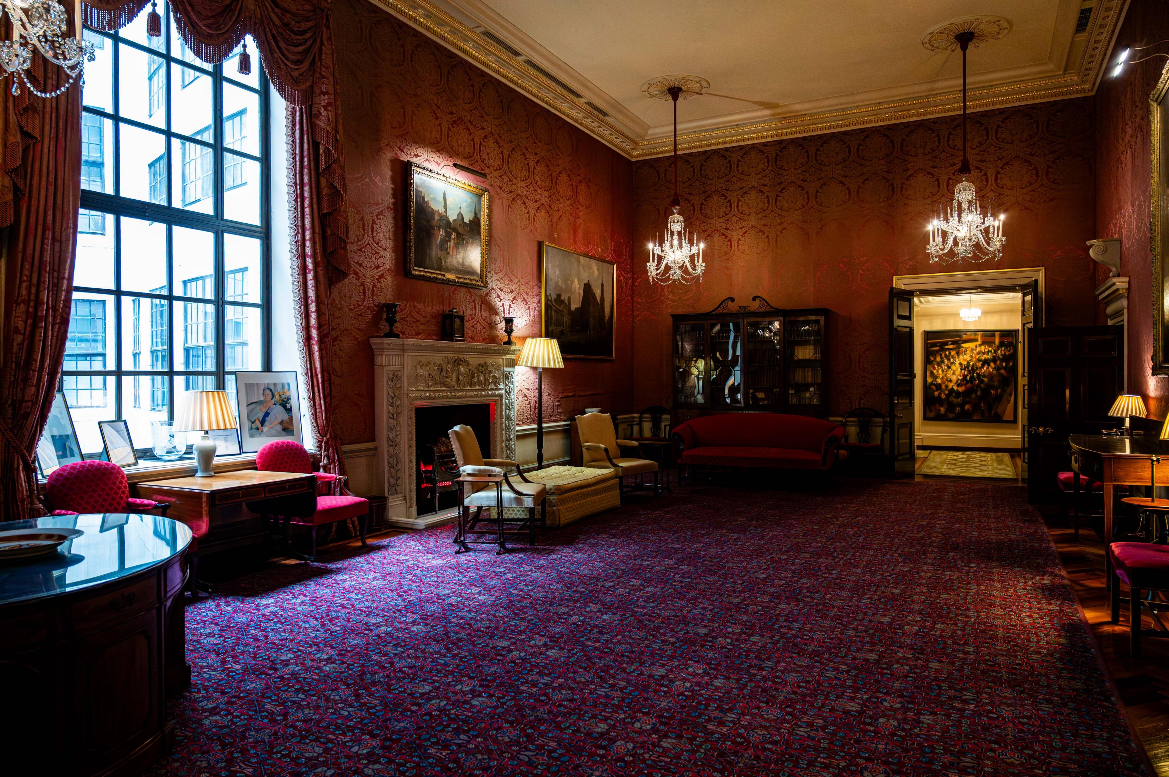 Ante room in the Bank of England which has red and gold silk-covered walls, chandeliers hanging from the ceiling and a blue, red and purple patterned rug on the floor. Pictures hang on the wall and there is a decorative stone fireplace, with other furniture around the room