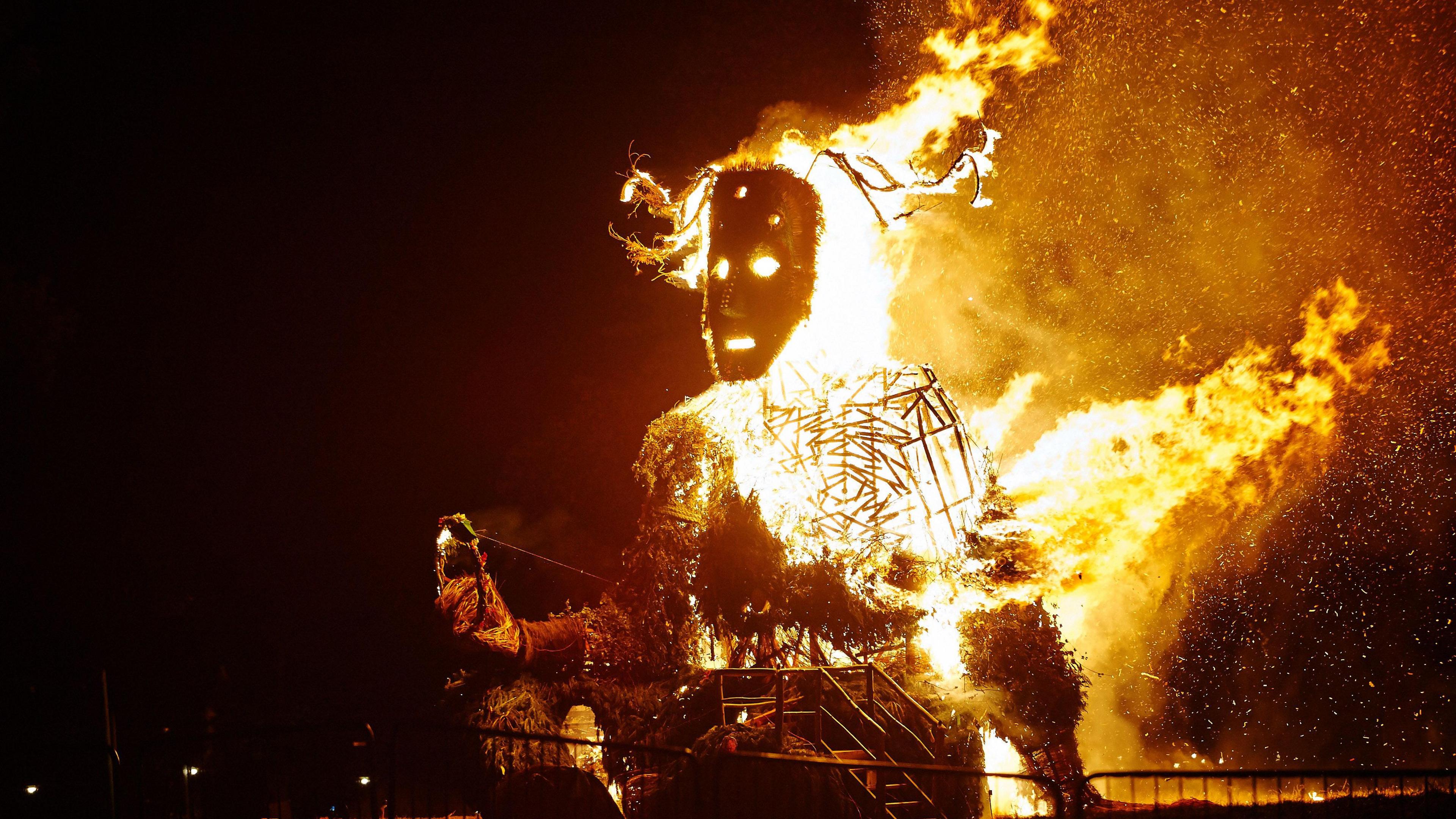 A giant Green Man sculpture has been lit on fire in the dark. Its face is illuminated with large flames coming from its torso. 