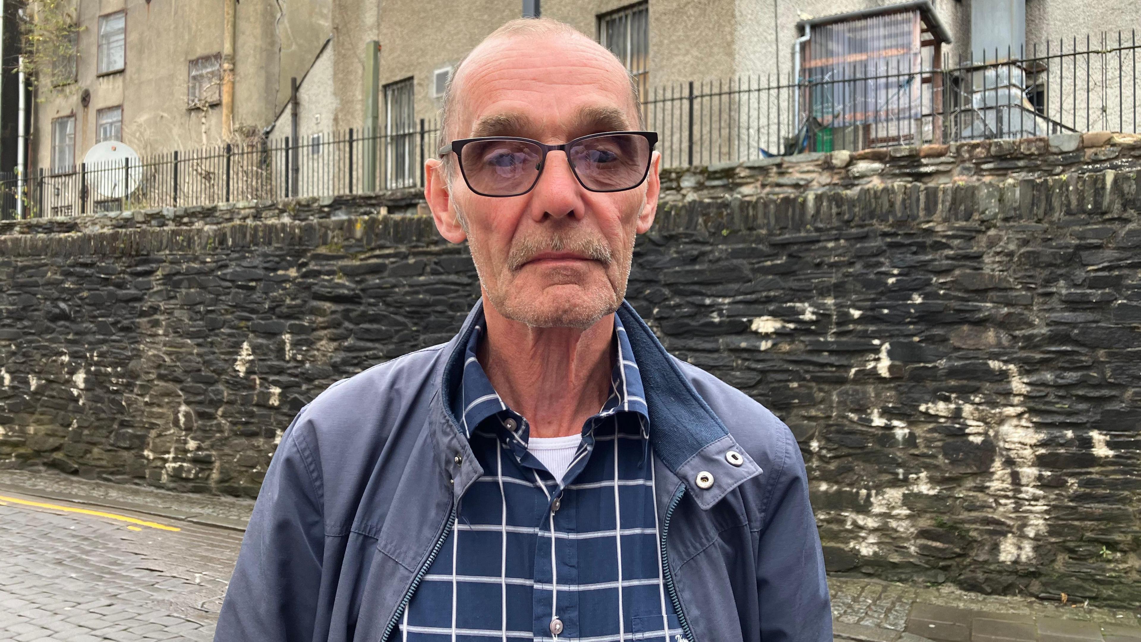 Liam Wray stands, serious faced, in front of the walls in Derry. He is weagin a blue jacket, white and blue checked shirt, and eye glasses.