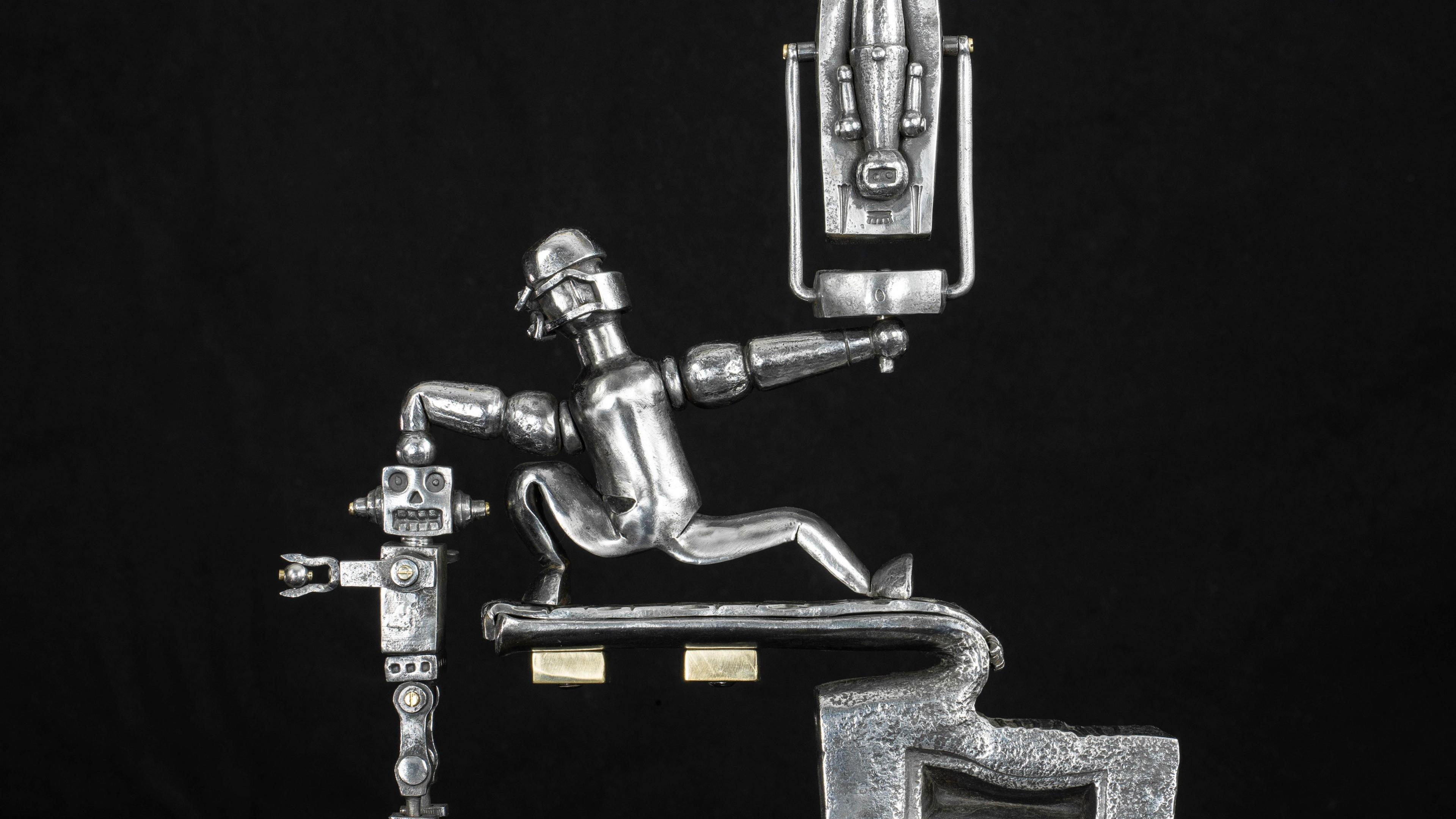 A silver metal sculpture shows a human figure standing astride a plinth, holding an indiscernible figurine up in his right hand and punching down on what looks like a robot with his left.