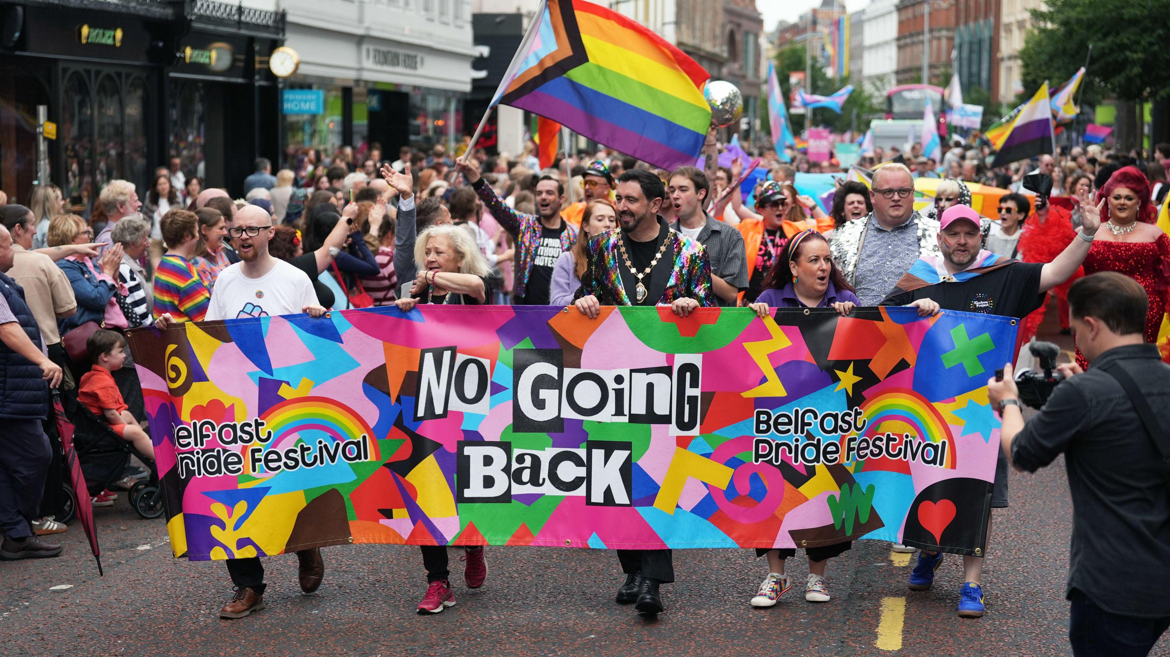 A crowd of people are marching through a city centre. A banner says no going back, Belfast Pride festival. Several people are waving flags.