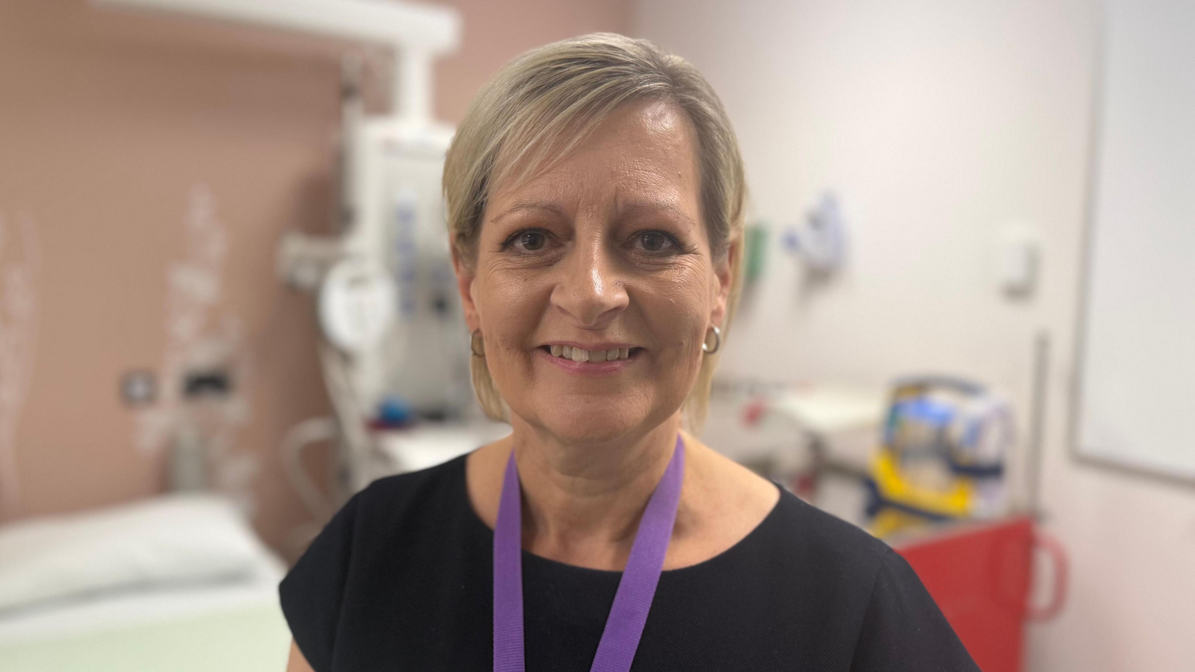 A lady with a blonde bob is smiling at the camera. She is wearing a black dress and a purple lanyard. She is standing in a hospital ward.