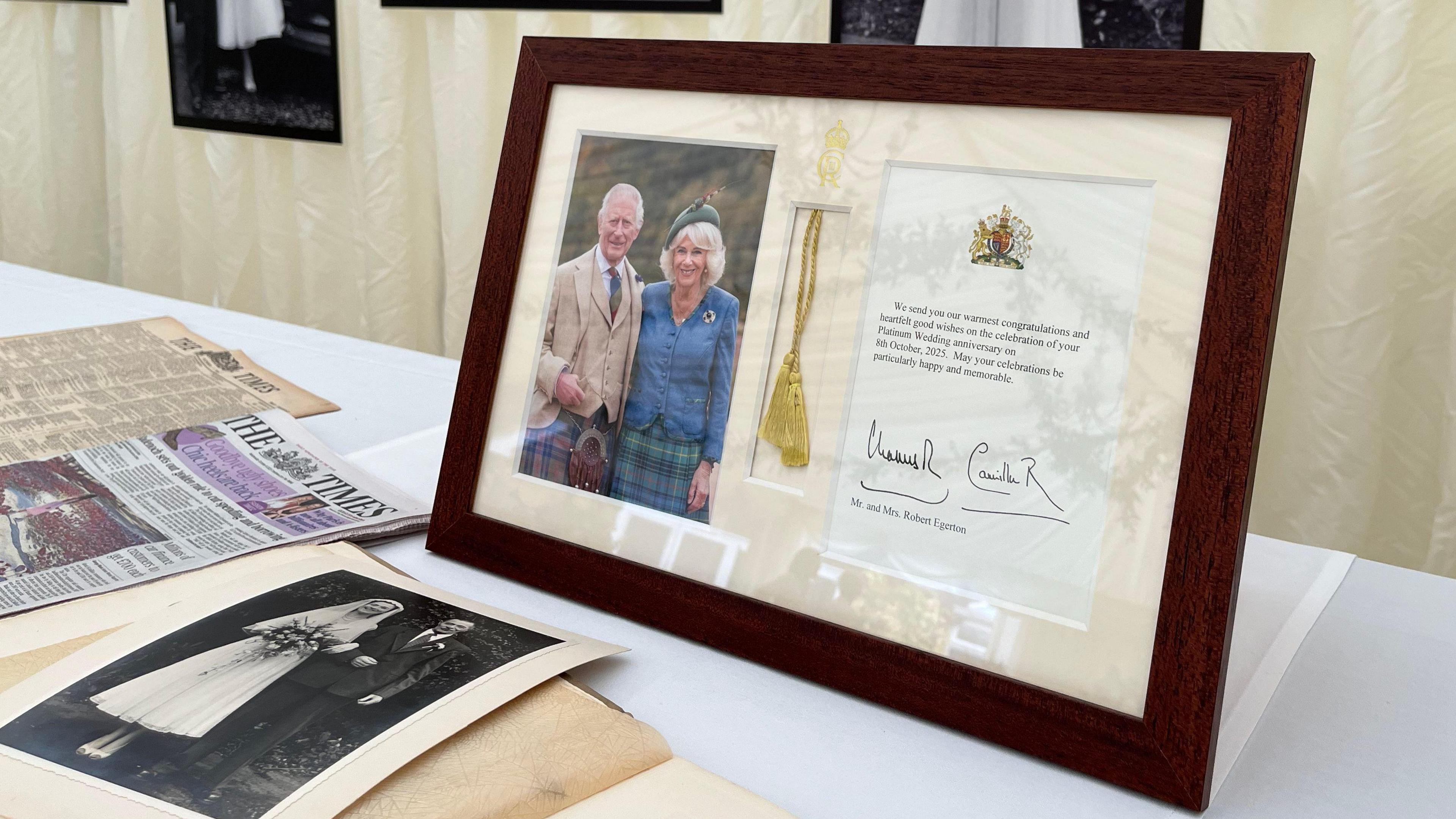 Framed message of congratulations from King Charles III and Queen Camilla, with copies of The Times newspaper from 8th October 1955 and 2025 in the background