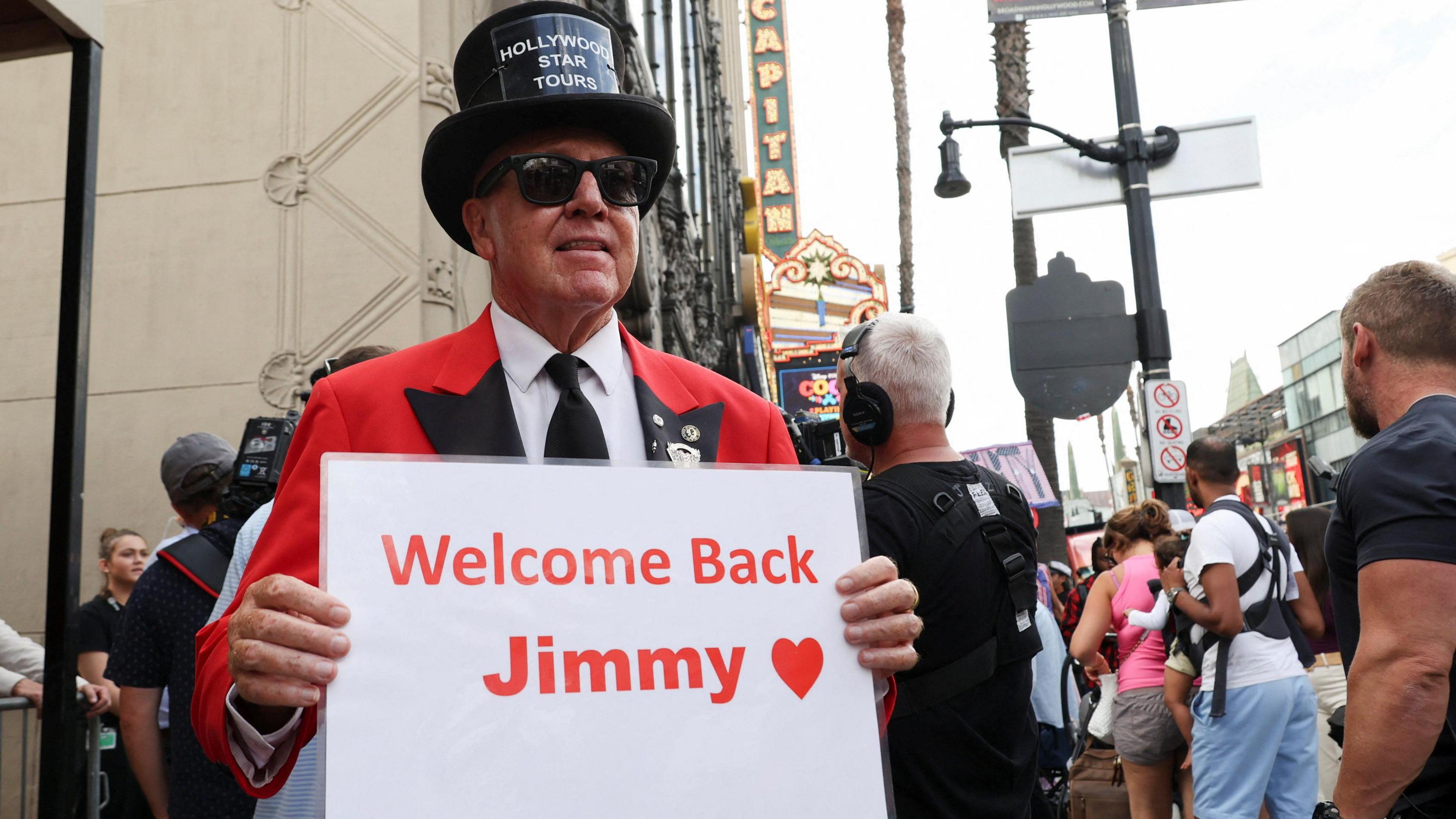 A man in a red suit and black top hat holding a sign saying "Welcome back Jimmy"