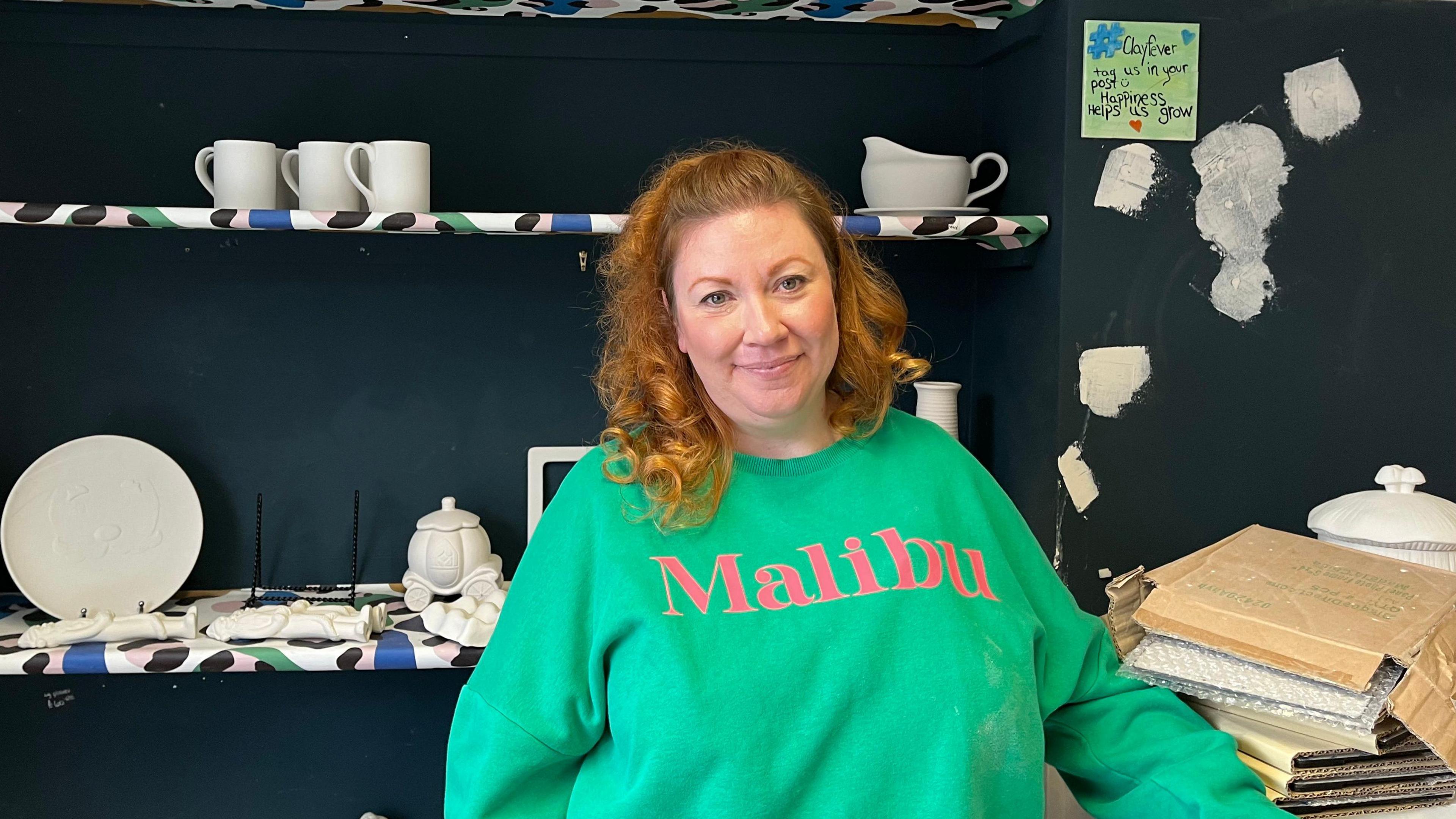 Woman with long ginger hair wearing a bright green jumper with the word "Malibu" across her front. She is stood in front of a dark blue painted wall with three shelves behind her. White plates, tea pots and cups are on the shelves. 