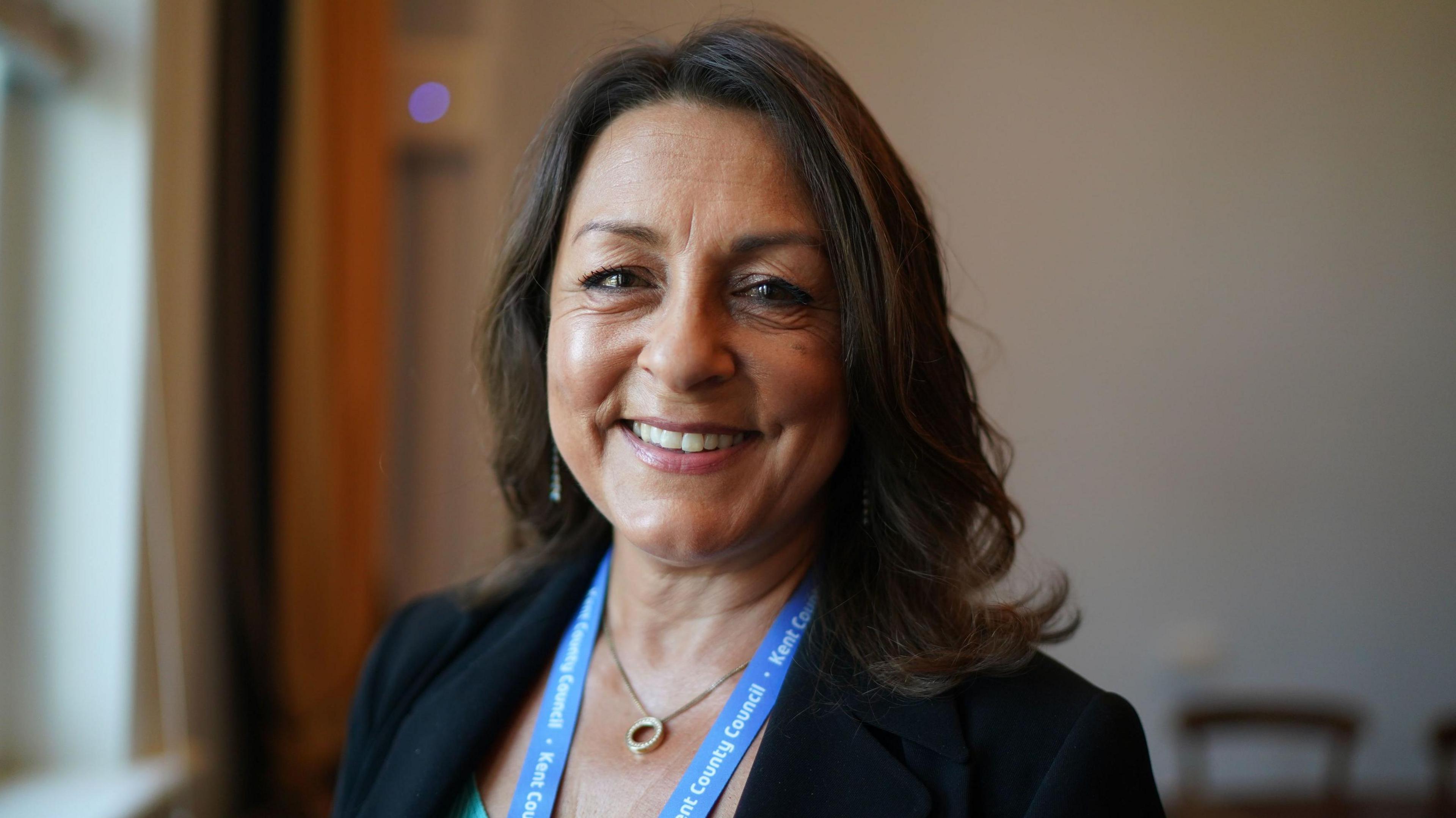 Linden Kemkaran, a middle-aged woman with long dark hair, wears a dark jacket and turquoise top, and has a turquoise Kent County Council lanyard around her neck. She smiles into the camera a she stands in an office.