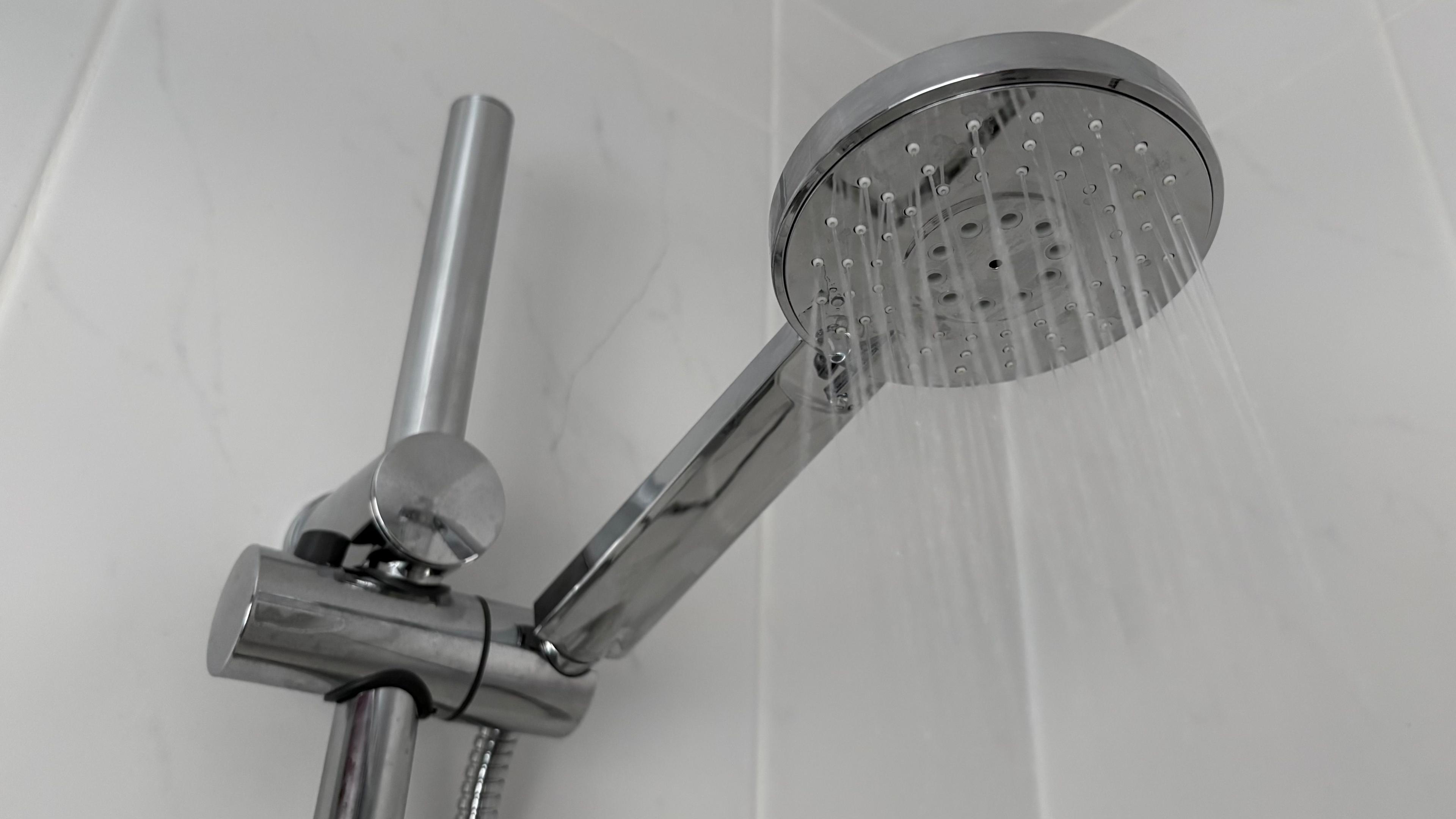 File shot of silver-coloured shower head with thin jets of water pouring out. It is held by an attachment which allows the height to be adjusted up a vertical pipe. The shower cubicle is covered with grey tiles.