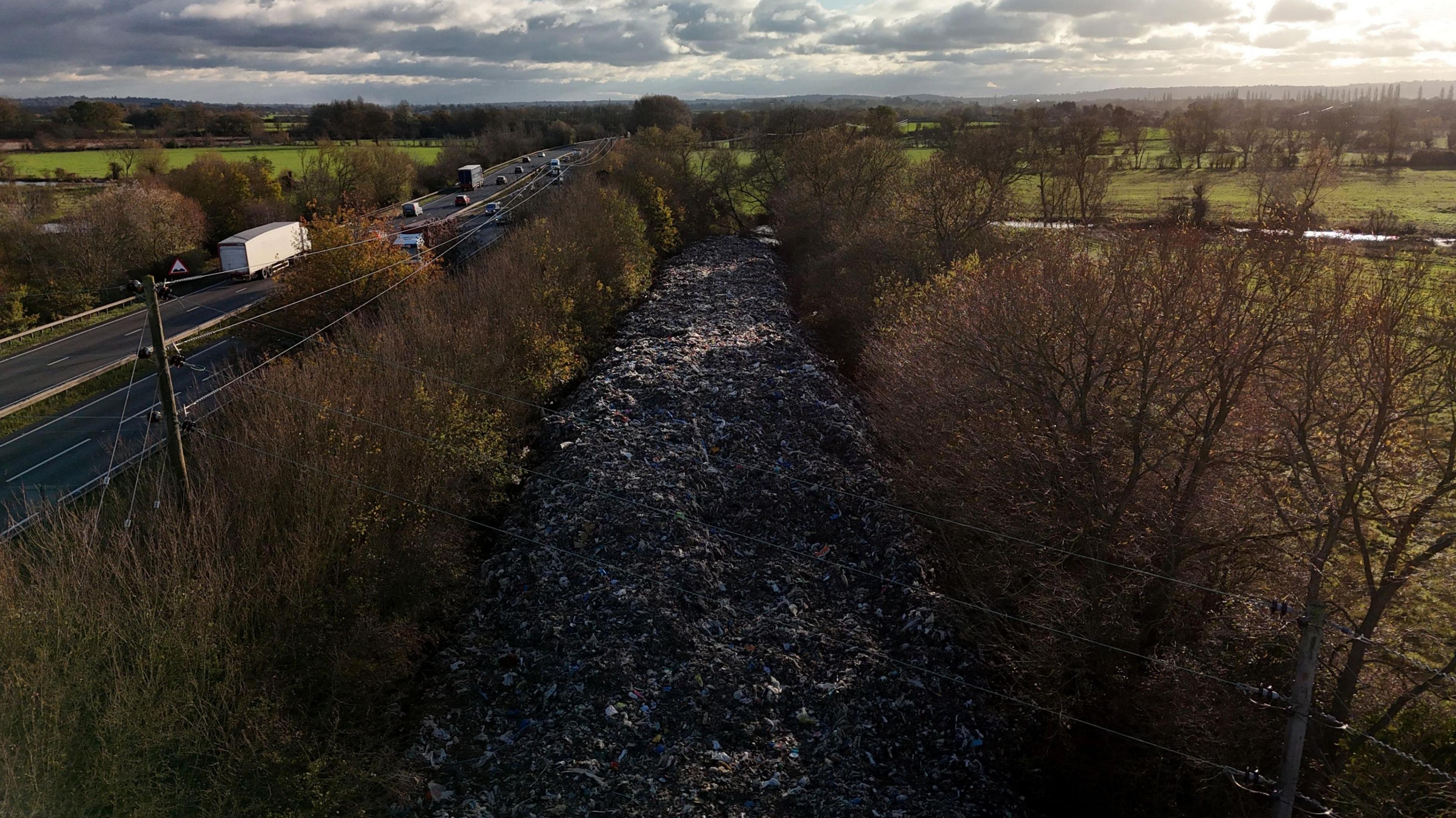 A huge pile of waste filling the length of a field between rows of trees, adjacent to a dual carriageway.