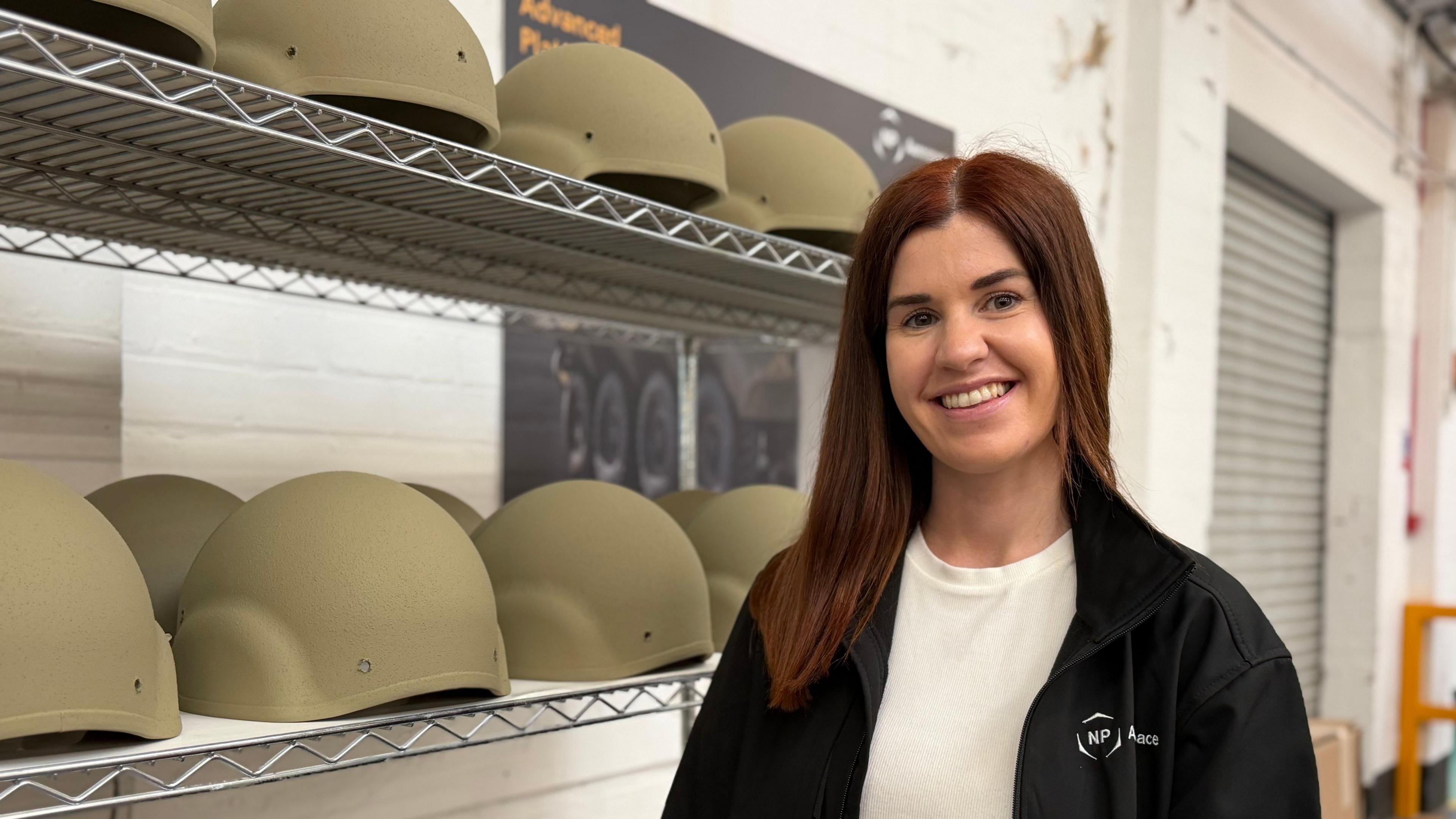 Erin Grieve stands next to wire racks filled with military helmets. She has long dark hair and is wearing a white T-shirt and black jacket.