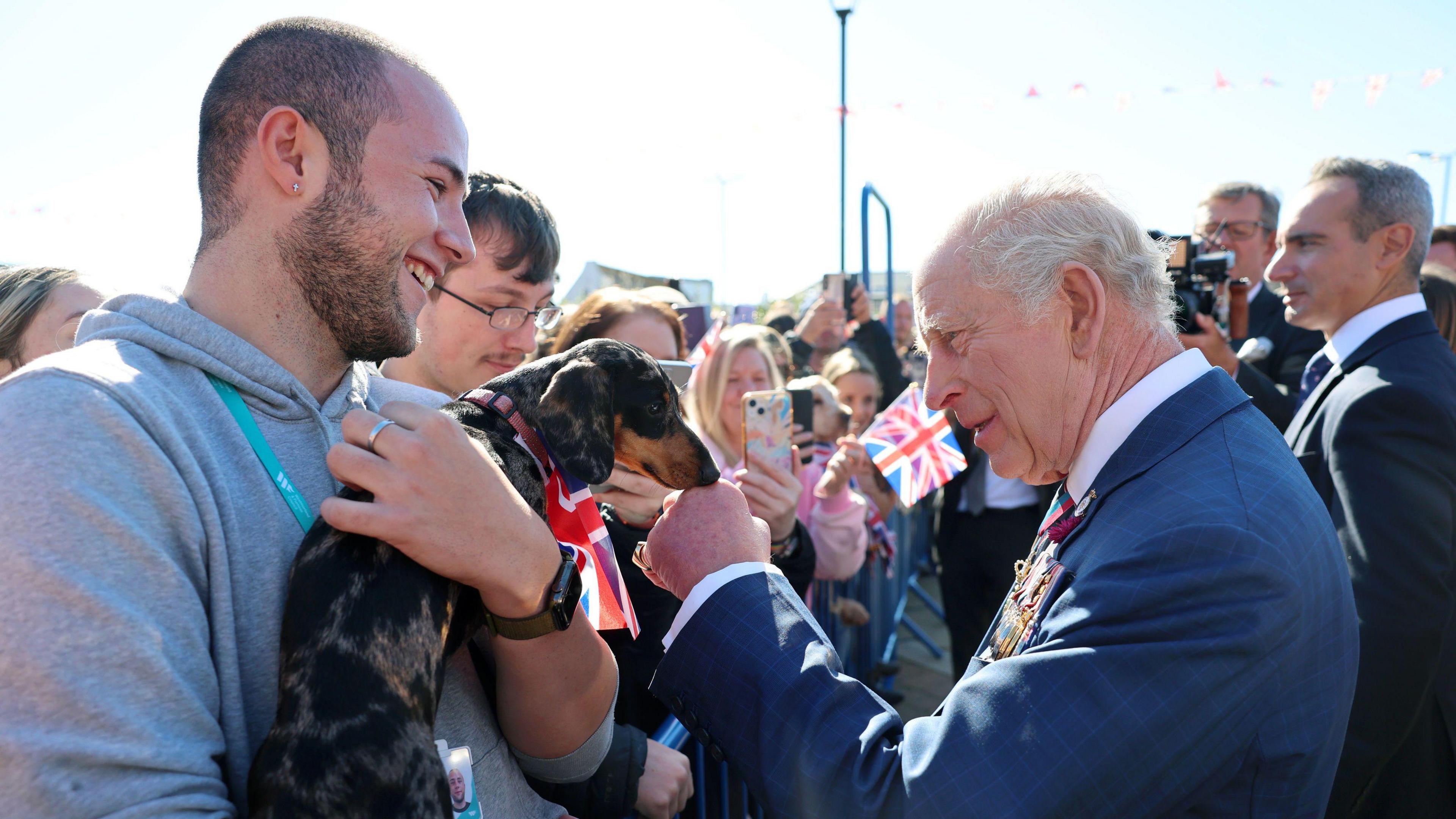 King Charles III strokes a dachshund as he greets members of the public. The dog has dark fur and has a union flag wrapped around its neck. It is being held by a man with short dark hair and a beard. He is wearing a grey hoodie.