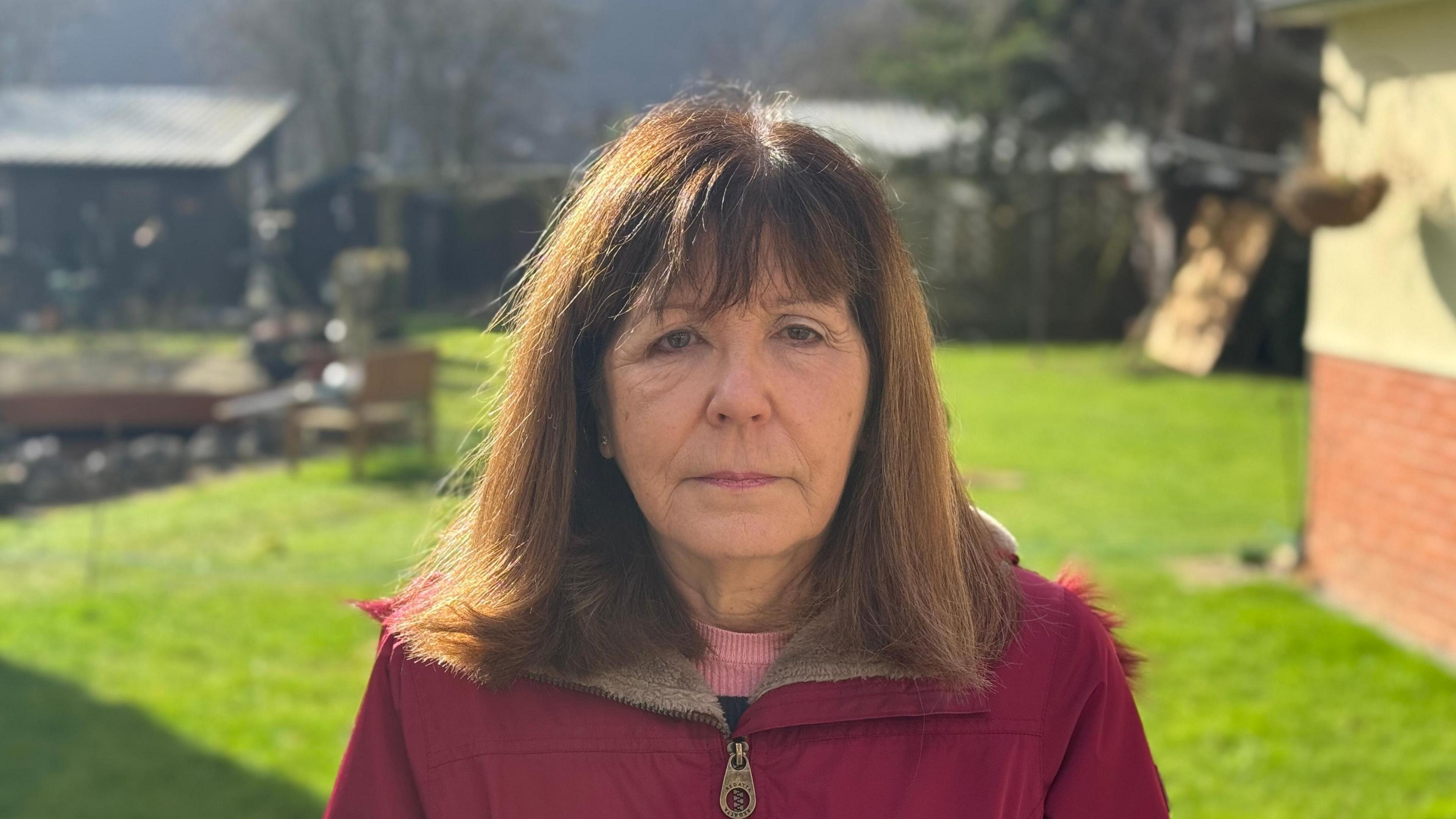 Sheila Snell - a woman wearing a red coat over a pink jumper standing in a back garden. The warehouse can be seen in the background.