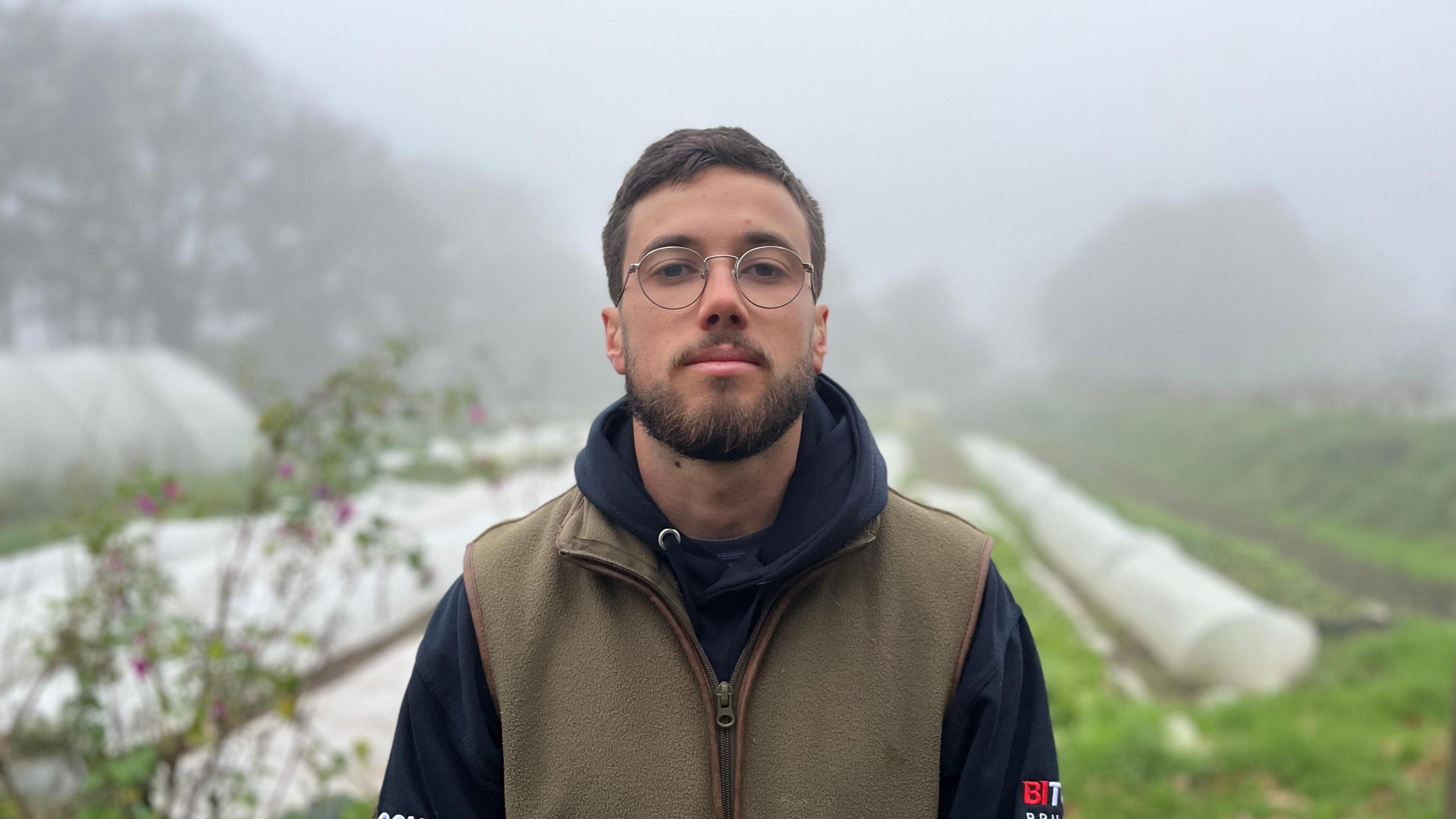 Oliver Griggs is in a man with short dark hair and a beard. He is wearing round glasses, a navy hoodie with a green, fleece bodywarmer on top. He is looking into the lens, with an out-of-focus misty field with plastic sheets on it in the background.