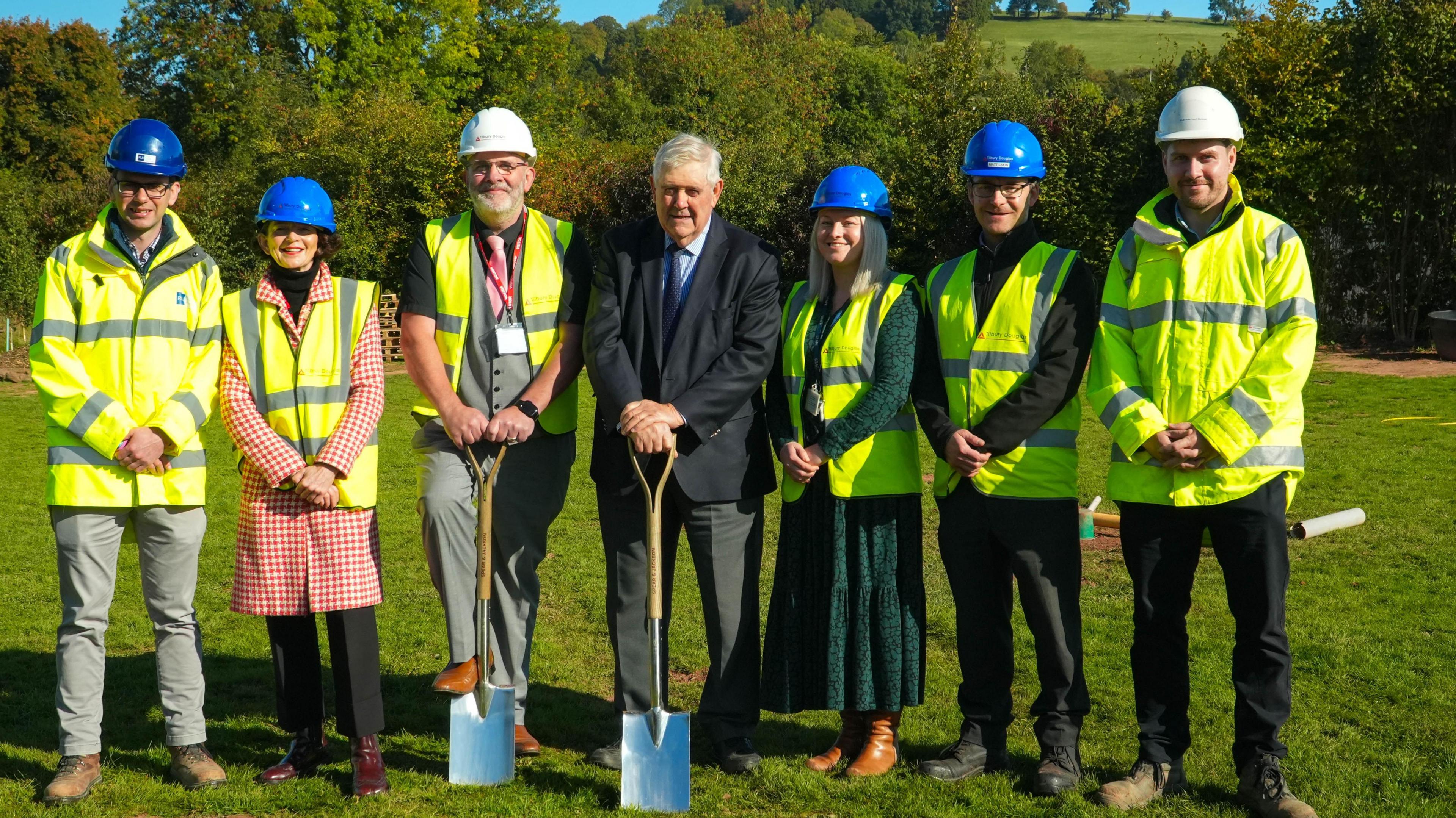 (Left to right) Chris Kenyon, Louise Tanner, Quentin Mee, Councillor Philip Price, Nia Powell, Matt Lakin and Daniel Horton stand with spades on the site. Most of them have hi-vis jackets and helmets. They are in a field with hills and trees behind them.