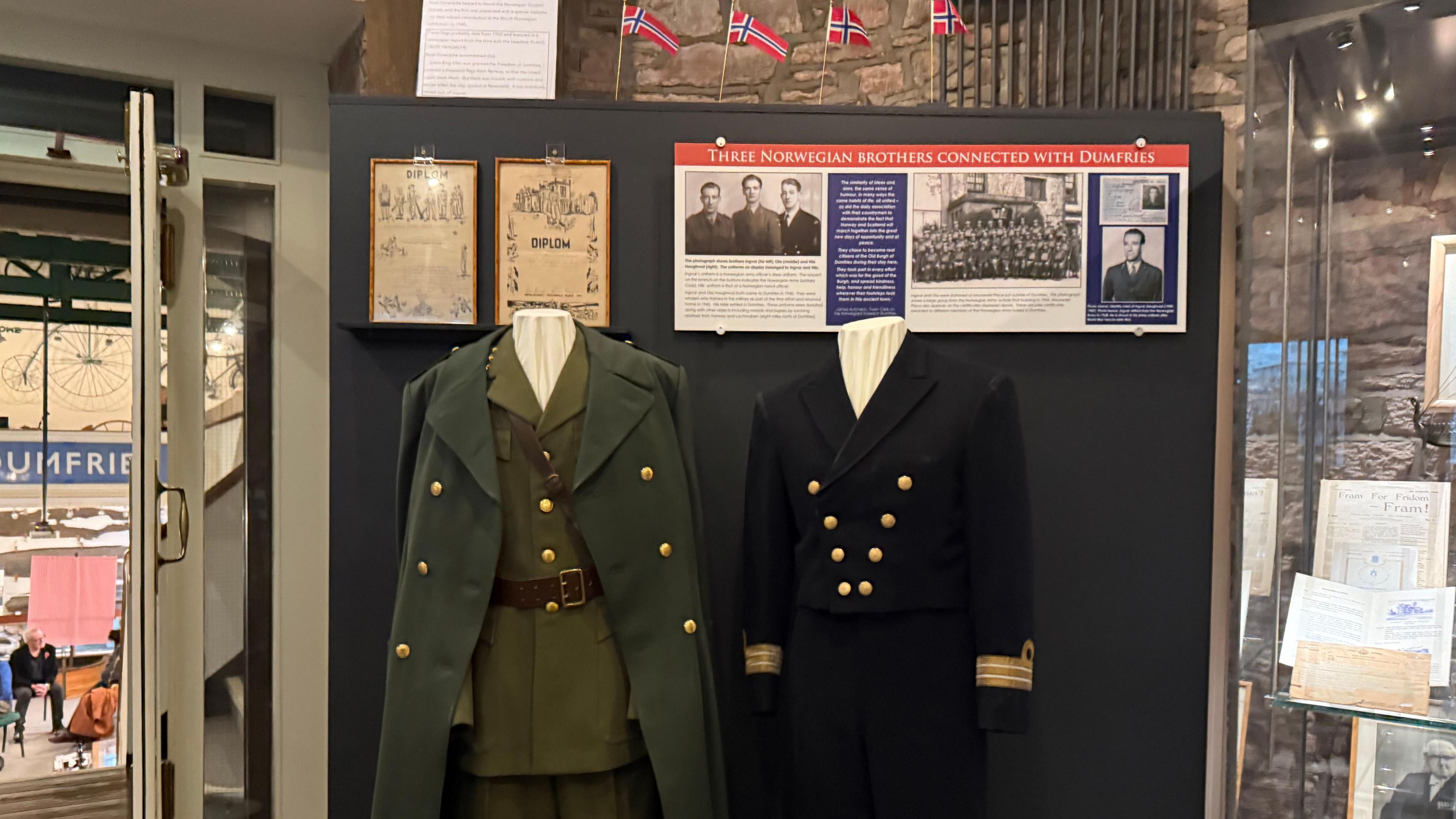 Two military uniforms positioned next to each other, green on the left, black on the right. They both have gold buttons.
